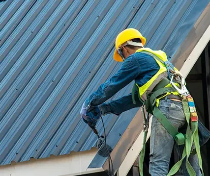 Worker wearing a yellow safety helmet and harness using a power tool on a metal roofing panel.