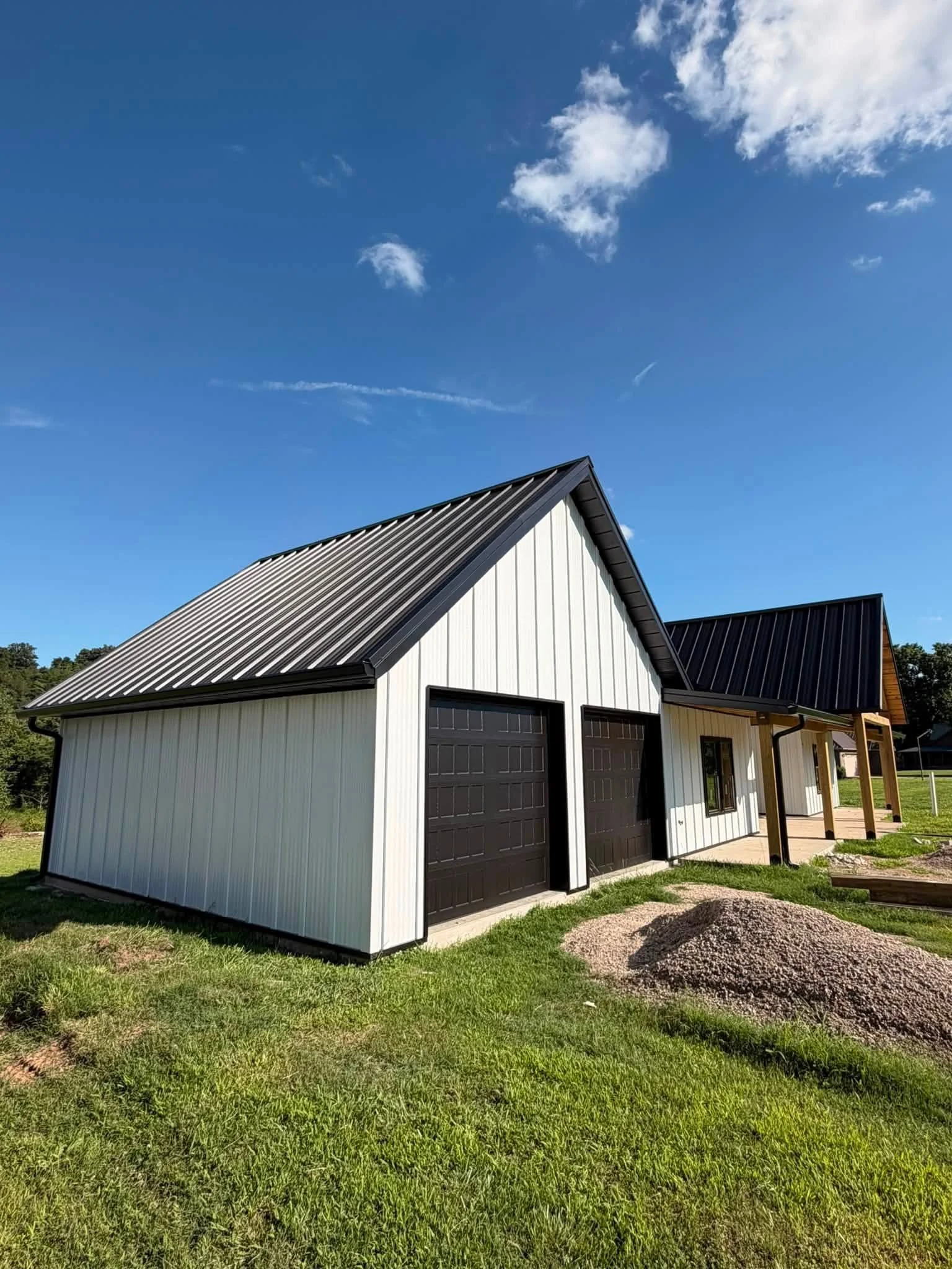 A modern barn-style building with black roof and white siding, two black garage doors, and a porch, surrounded by green grass and a dirt pile in the yard. The sky is blue with some white clouds.