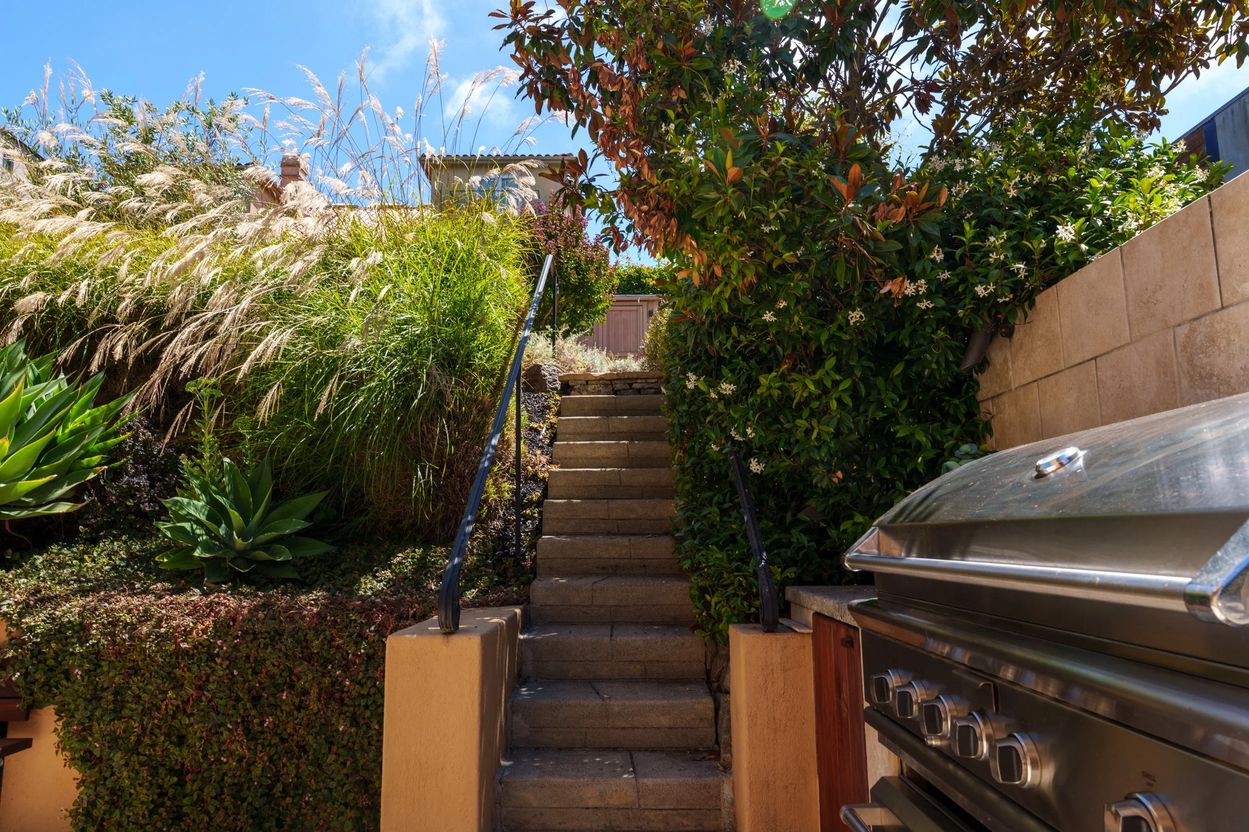 Outdoor staircase with concrete steps, black metal handrails, surrounded by lush green and flowering plants, leading up to a house with a tiled roof, under a clear blue sky.