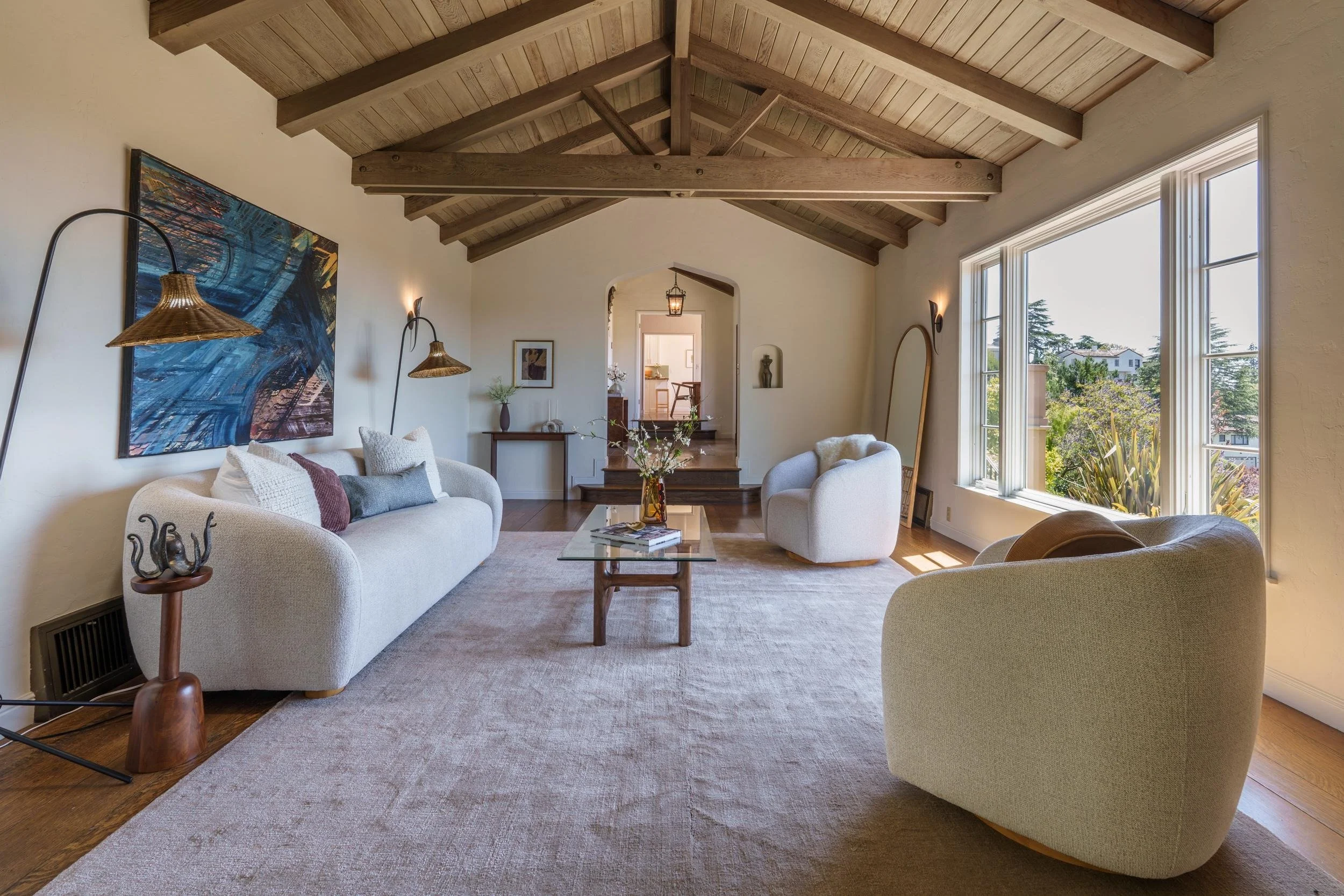 A cozy living room with a vaulted wooden ceiling, large windows, and white furniture including two armchairs and a sofa at a glass coffee table. Decorative items such as a large abstract painting, lamps, and a vase with flowers are visible.
