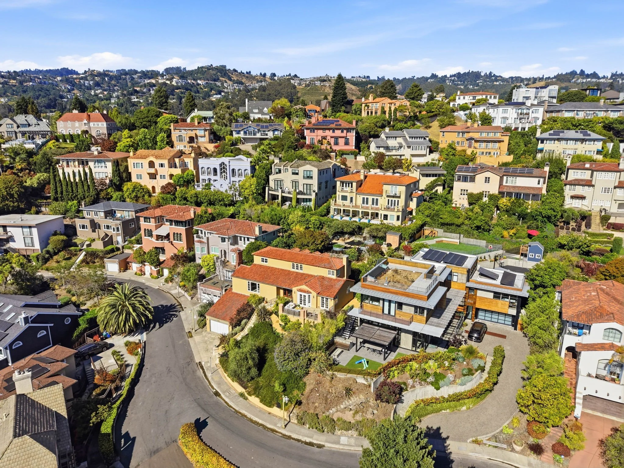 Aerial view of a hillside residential neighborhood with colorful houses, trees, and a winding street under a blue sky.