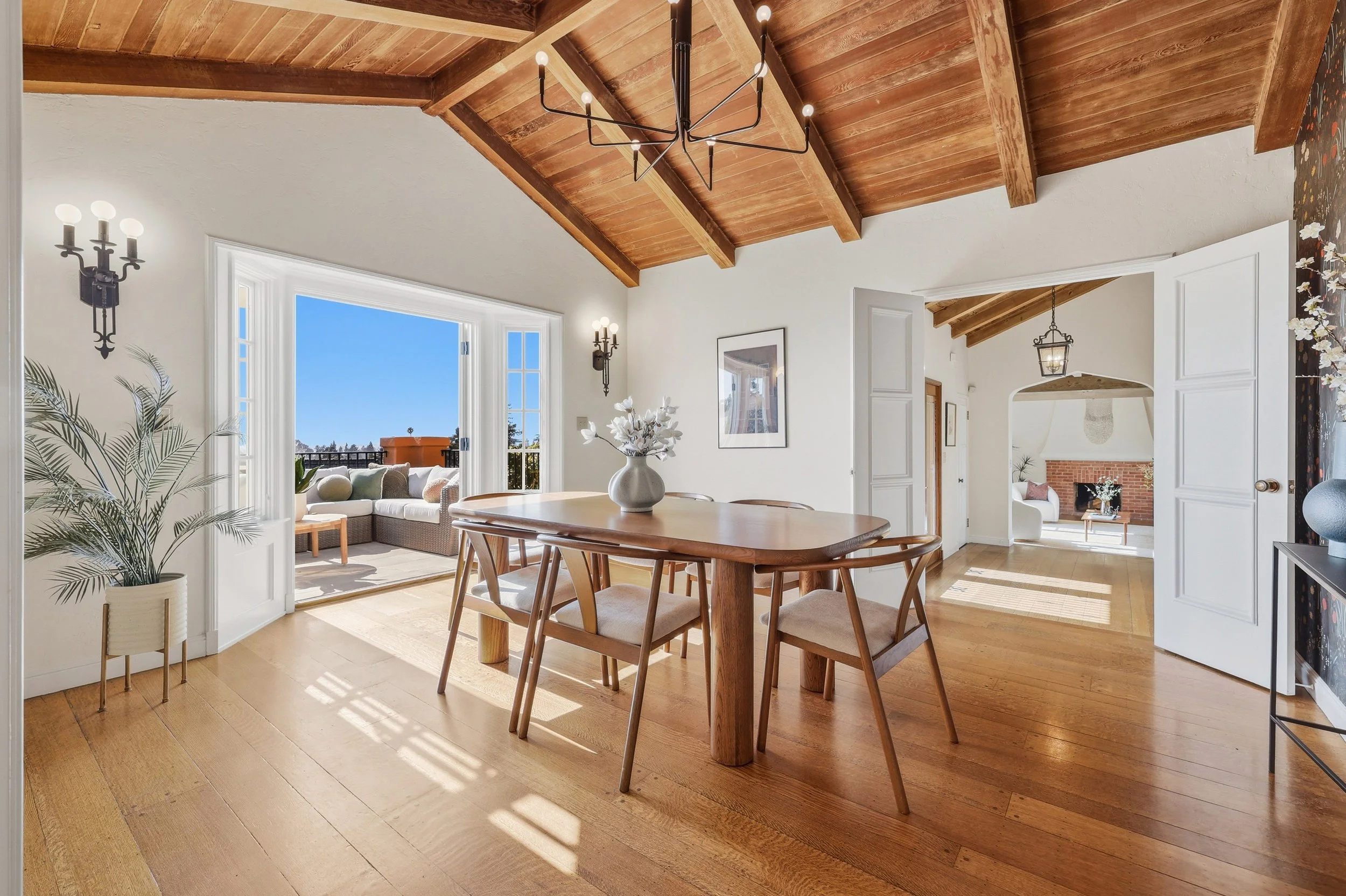 Dining room with wooden table, chairs, large vase with white flowers, a wall hanging, and open balcony door showing outdoor seating and a view of trees and blue sky.