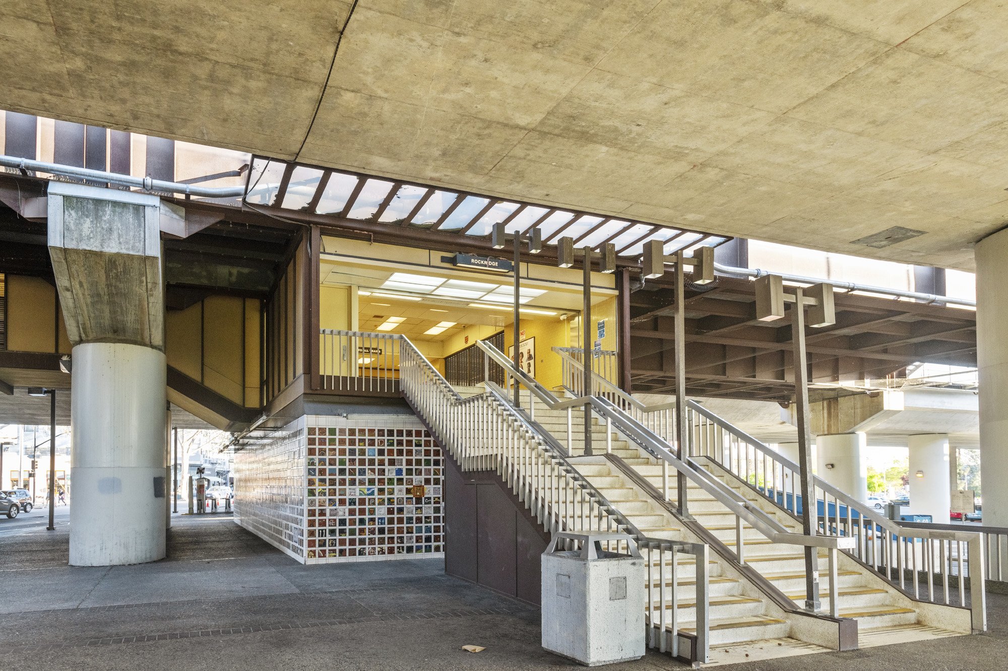 An outdoor stairway leading to an entrance of a building under an overpass, with a yellow painting and signs visible inside.