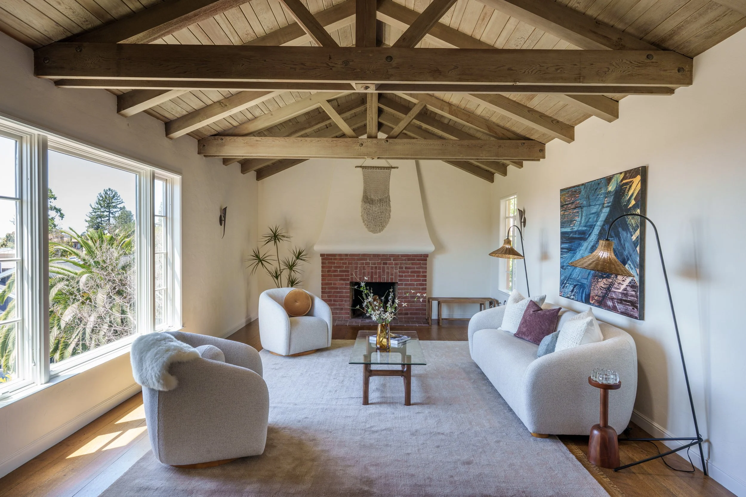 Living room with large windows, white sofa, armchair, coffee table, brick fireplace, artwork, and wooden ceiling beams.