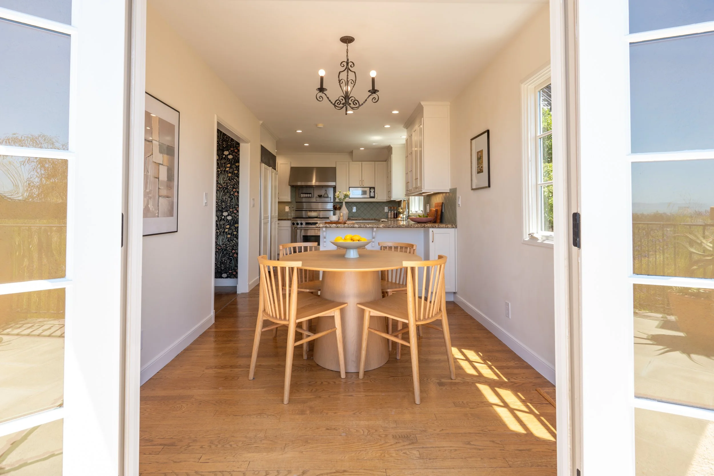 Bright kitchen and dining area viewed through open French doors, with a round wooden table and six chairs, a ceiling chandelier, white cabinetry, and windows bringing in natural light.