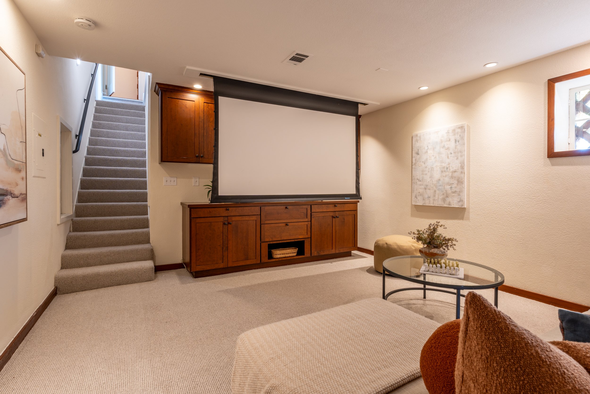Living room with a large projector screen, wooden cabinet, staircase, and artwork on textured walls.