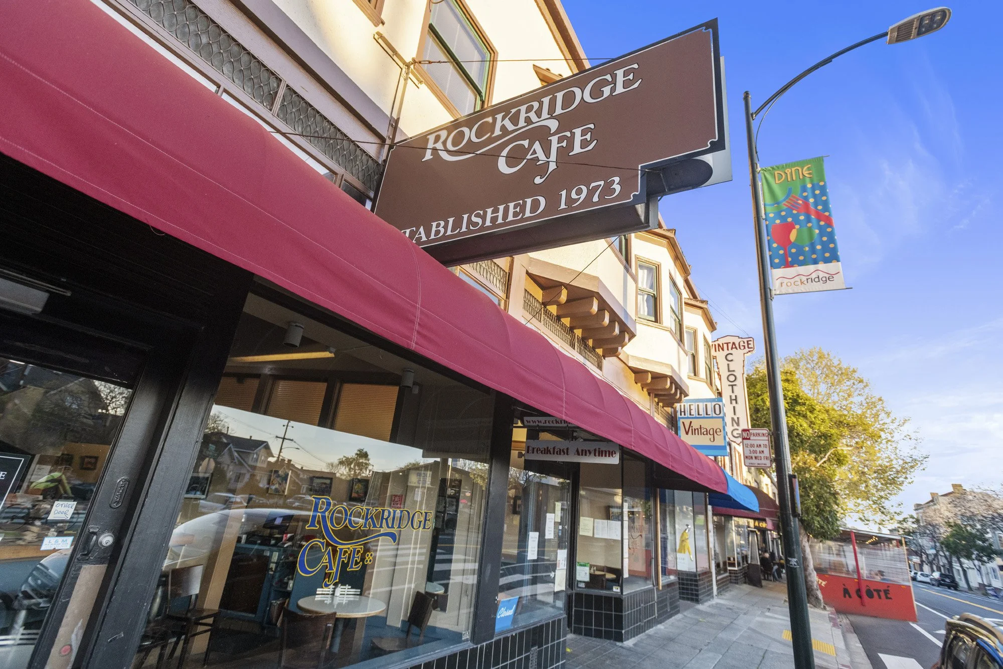 Street view of the Rockridge Cafe, established in 1973, with a red awning and a large brown sign. There are colorful banners and signs advertising dine-in, breakfast any time, and vintage clothing shops nearby.