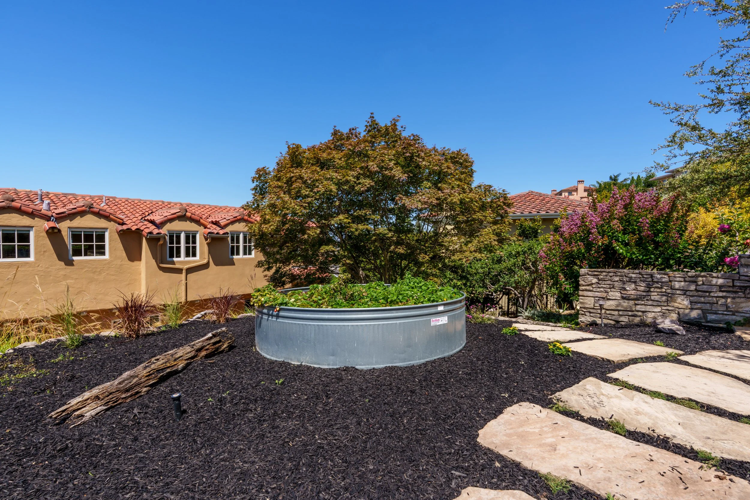 A backyard garden with a circular raised planter filled with green plants, a weathered log laying across dark mulch, a stone pathway, a large tree with reddish leaves, pink flowering bushes, and a house with a red tile roof and beige walls under a cl
