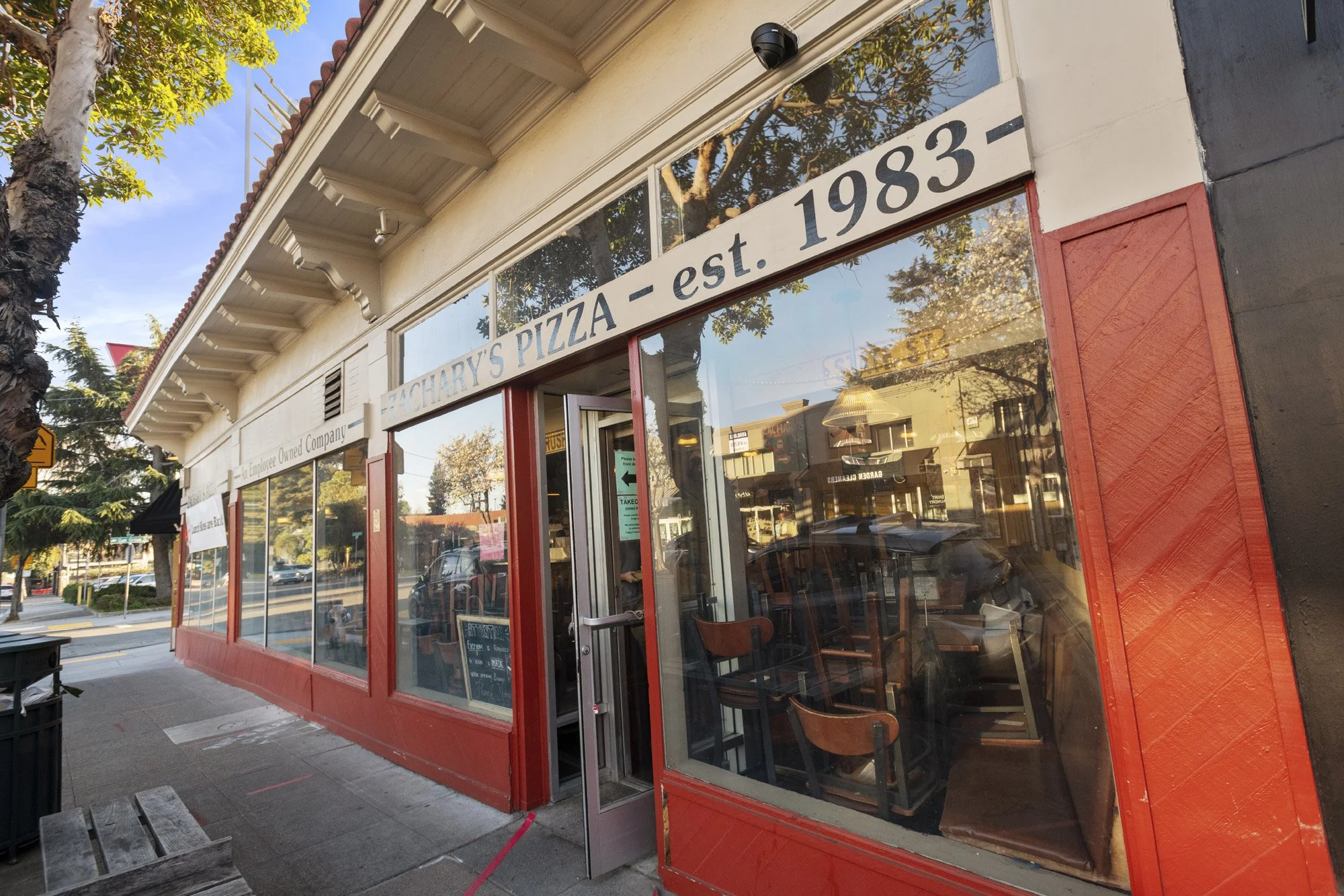 Front view of Zachary's Pizza restaurant with large windows and red trim, established in 1983, outdoor seating area, and trees reflecting in the windows.