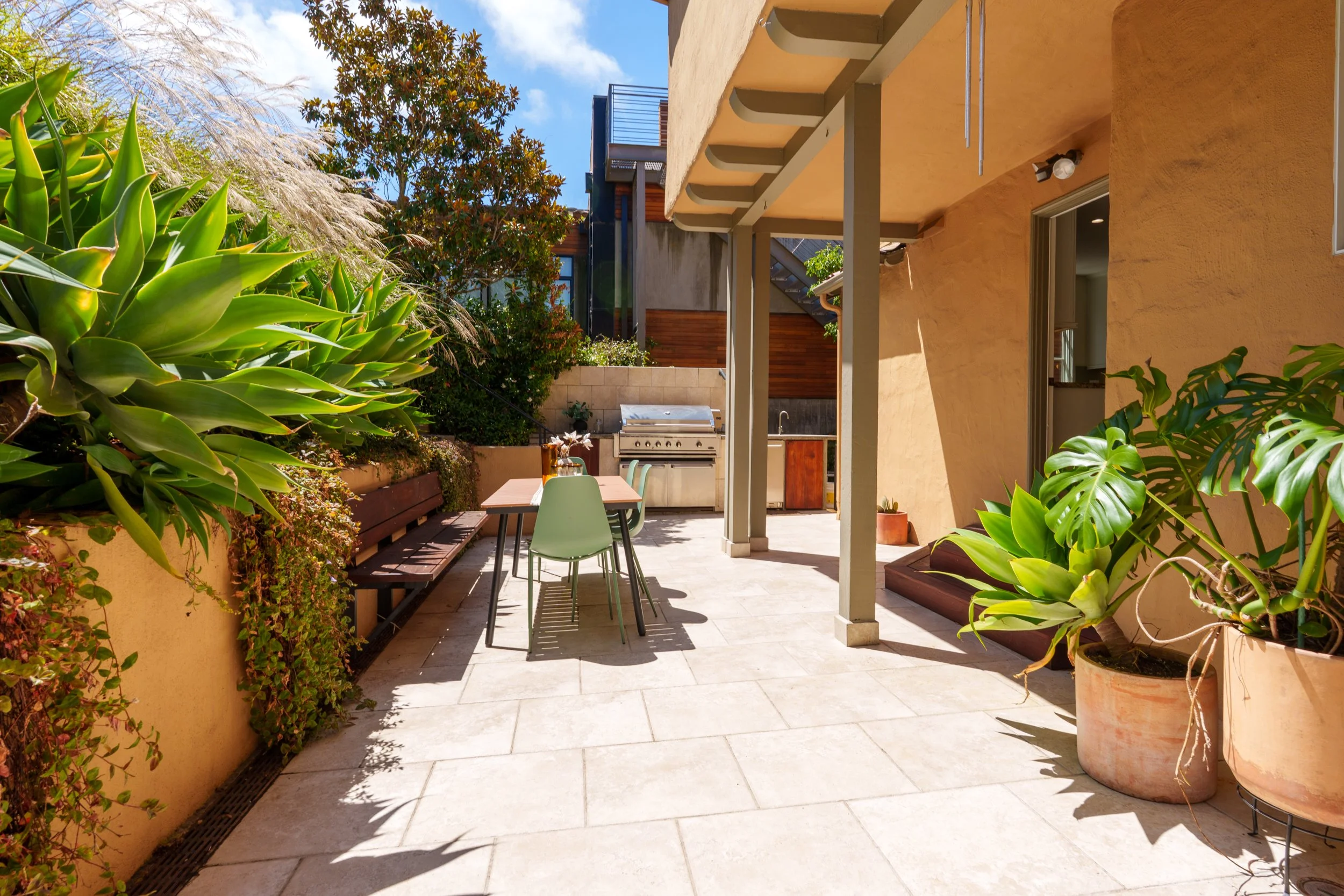 Outdoor patio with large green potted plants, a table with chairs, built-in grill, and yellow stucco walls under a sunny sky.