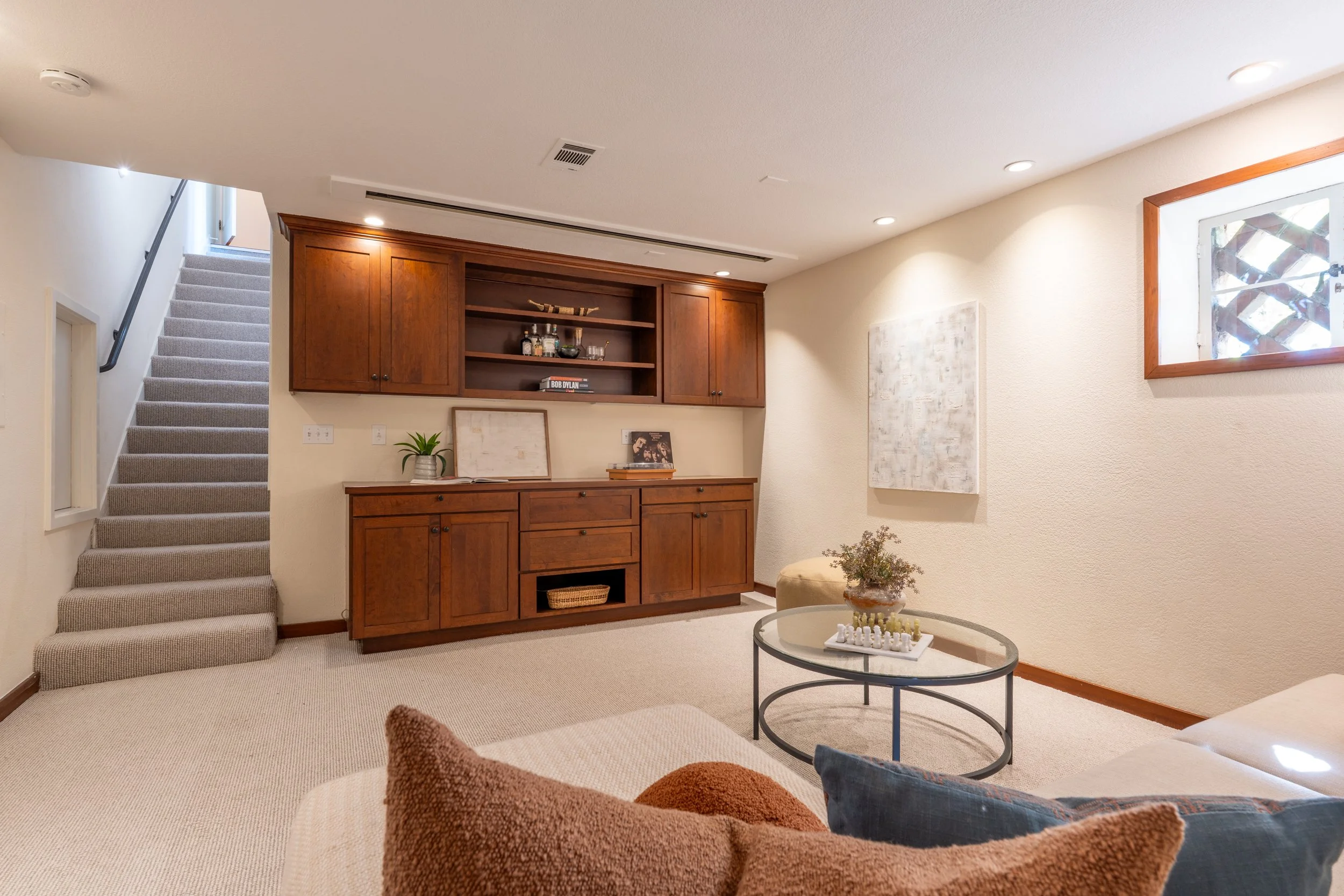 Living room with beige carpet, wooden wall unit with open shelves, staircase with carpeted steps, round glass coffee table with chess set, painting on wall, window with wooden frame, and a cream-colored sectional sofa with cushions.