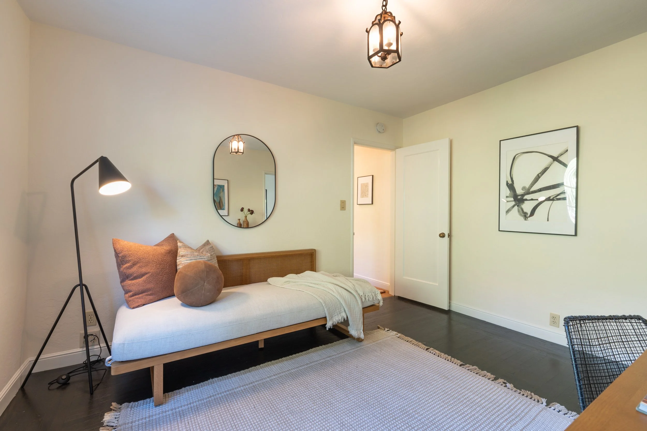 A bedroom with white walls, a bed with beige and brown pillows, a black floor lamp, and artwork on the walls. There is a door slightly open, revealing another piece of framed art. A light fixture hangs from the ceiling, and a white area rug covers pa