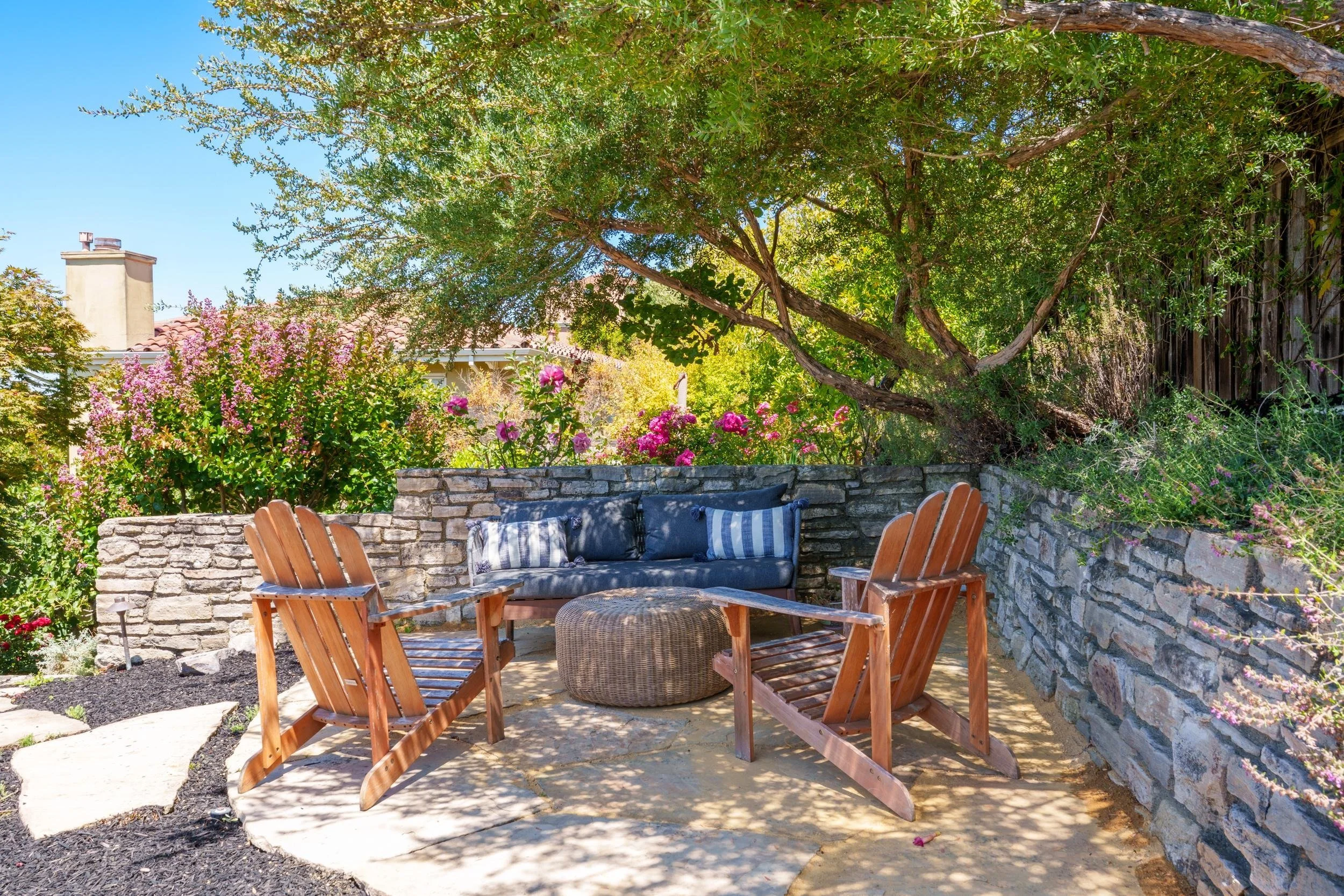 A cozy outdoor patio area with a stone wall, wooden chairs, and a cushioned outdoor sofa surrounded by lush greenery and colorful flowers.