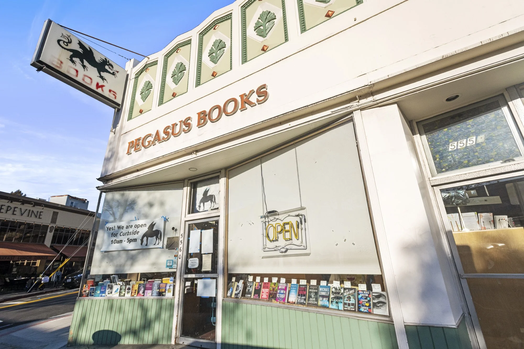 Front view of Pegasus Books store with a sign showing a black horse silhouette, large windows displaying books, and an open sign on the door. The storefront has a white exterior with green accents.