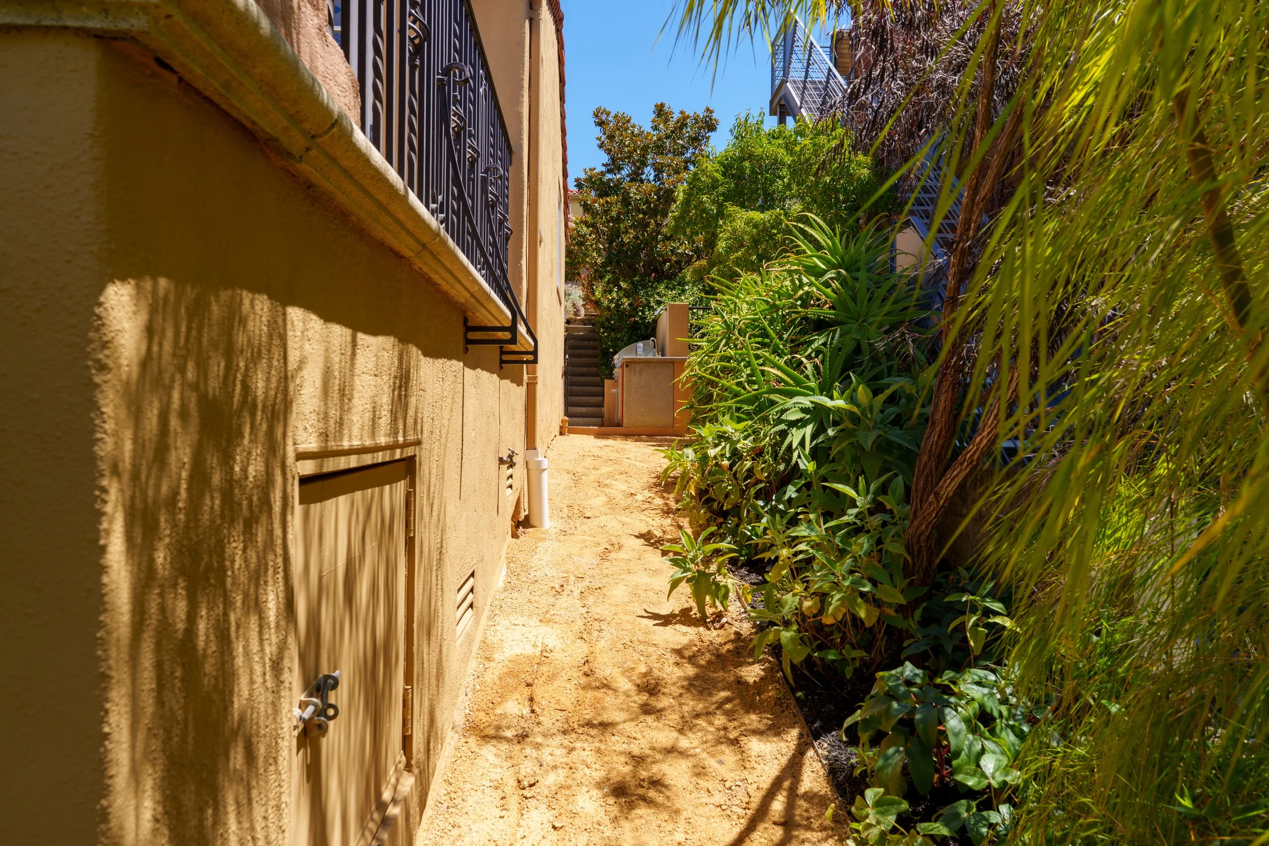 Narrow outdoor pathway between a beige stucco building with decorative metal window bars and lush green plants on the right, with a clear blue sky above.