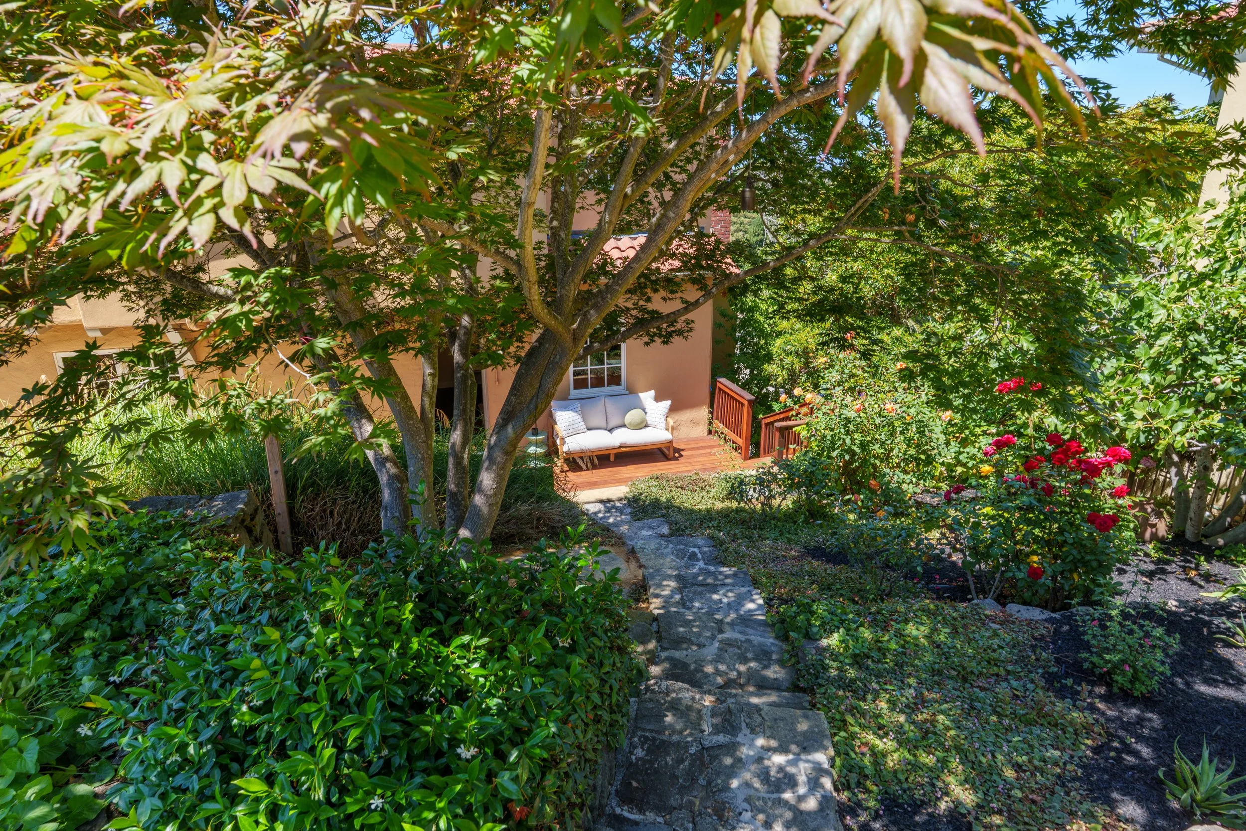 A lush garden with a pathway leading to a house, surrounded by trees, flowers, and greenery, with outdoor seating on a wooden deck.