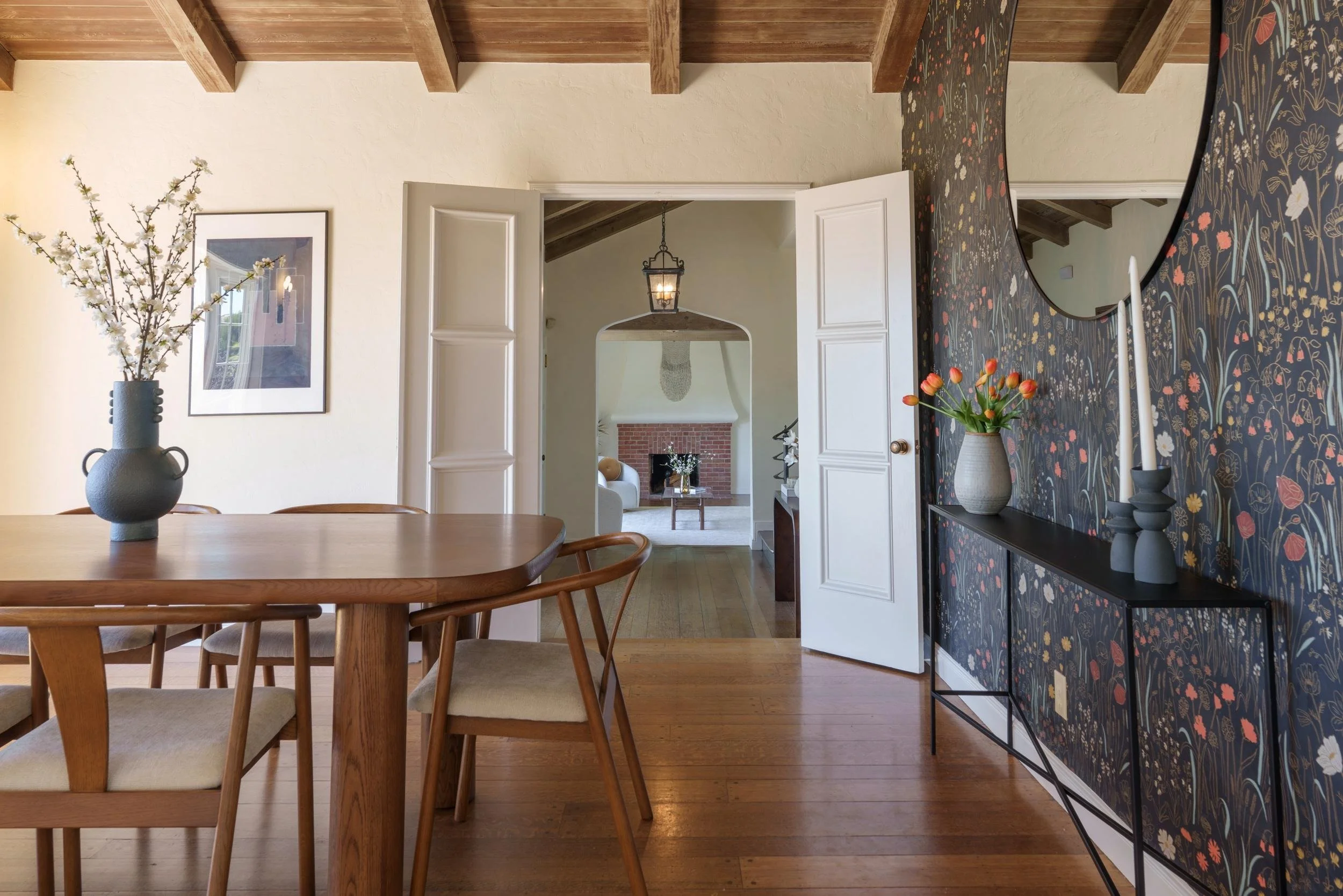 A dining room with a wooden table, chairs, and decorative vases with flowers. An open door leads to a living room with a fireplace, and a mirror on the wall reflects part of the room.