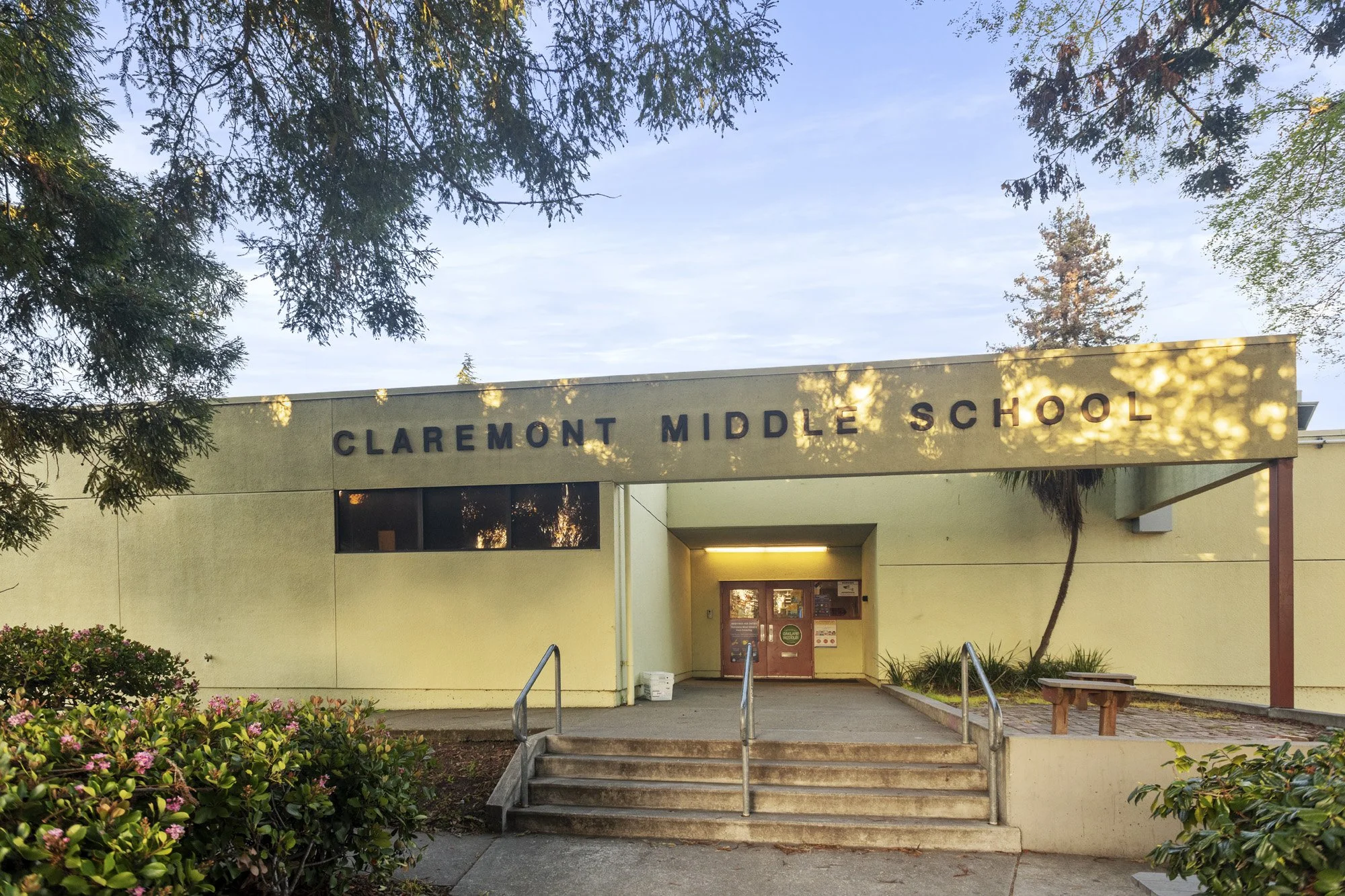 Photo of Claremont Middle School entrance with a sign, steps leading up to the door, surrounded by trees and bushes.
