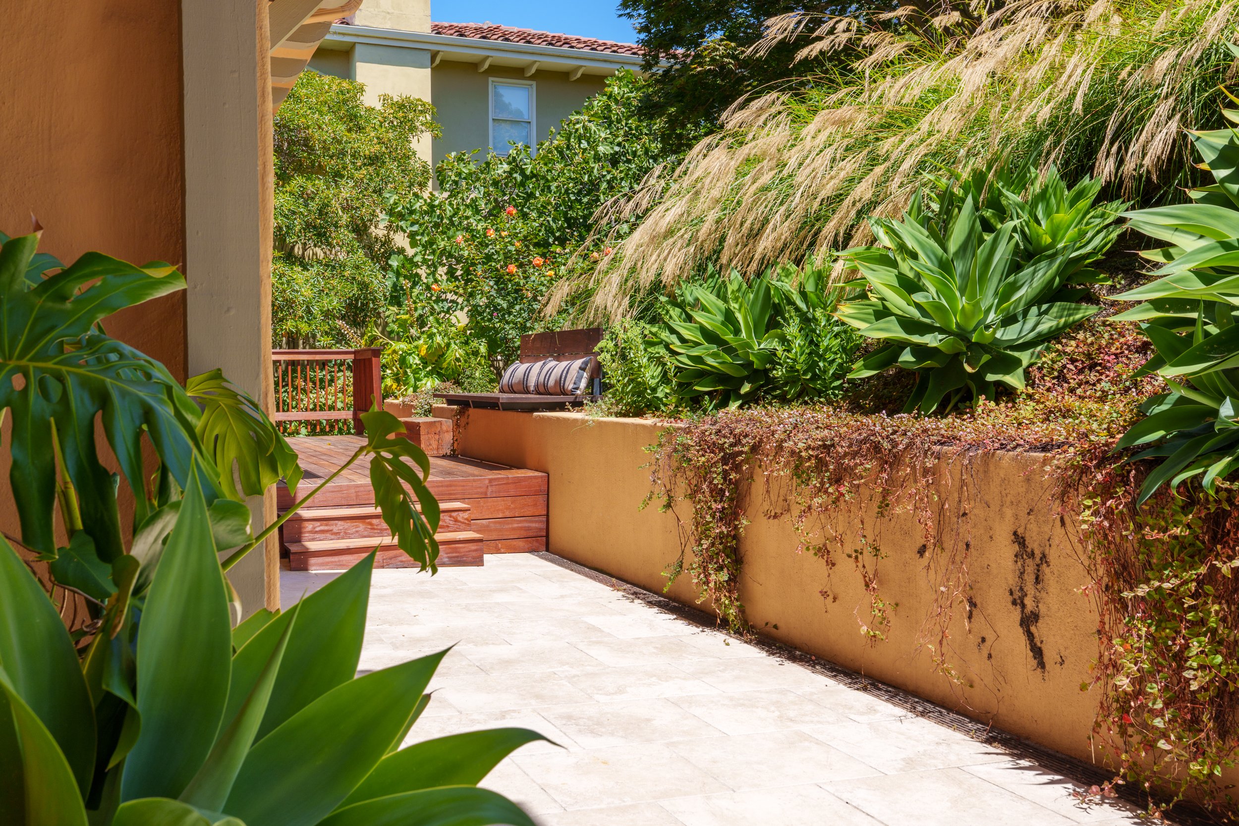 A cozy backyard patio with lush green plants, tall grasses, and flowering bushes, featuring a wooden bench with a striped cushion, surrounded by warm-colored walls and a beige tiled floor under a sunny sky.