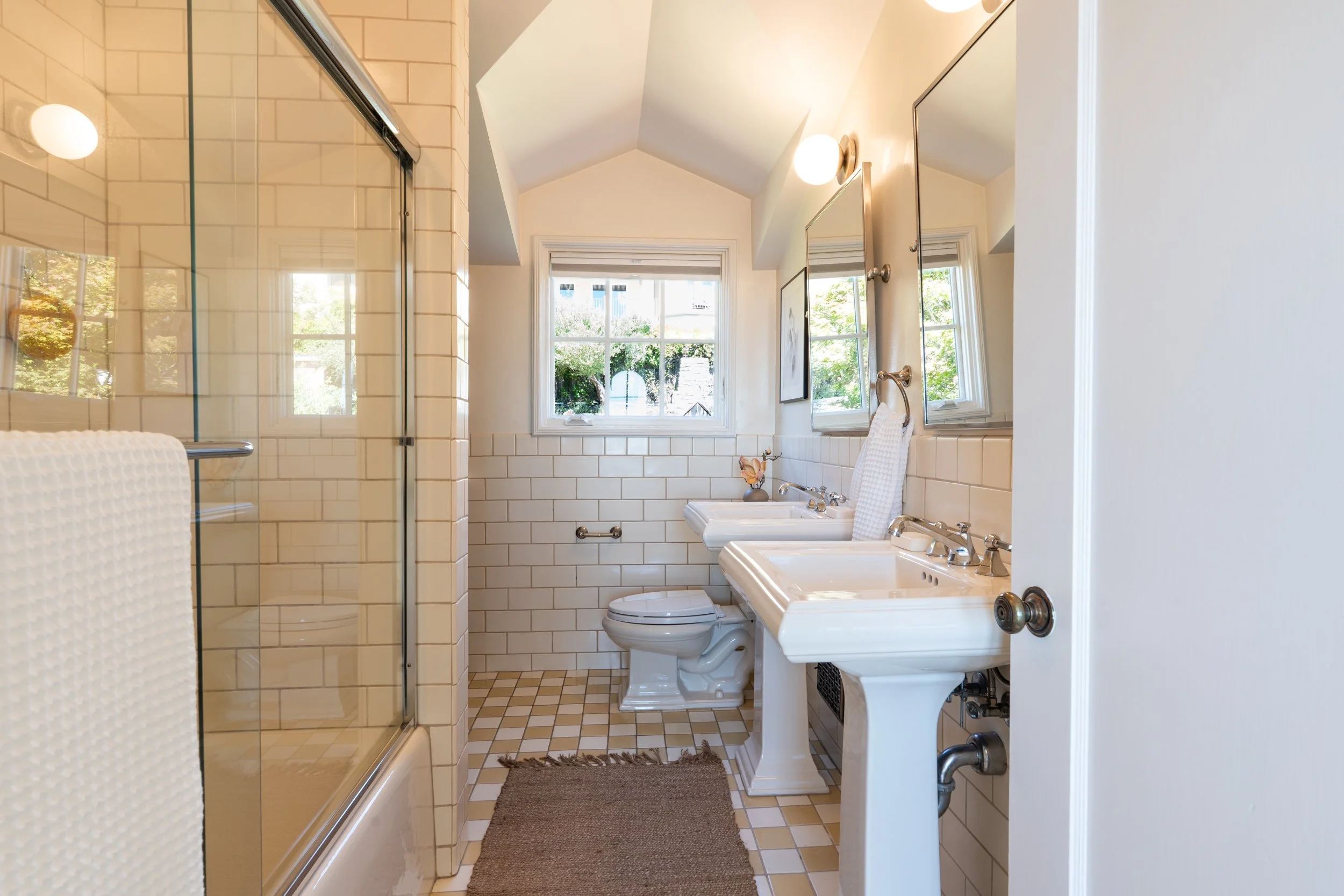 A bathroom with a tiled shower area, a window letting in natural light, two sinks with mirrors, a toilet, and a small rug on the tiled floor.