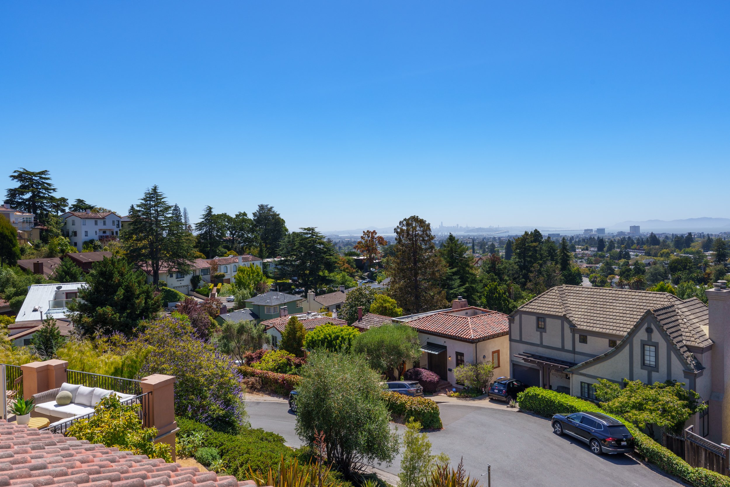 A view of a neighborhood with houses, trees, and cars, with a city skyline and mountains in the distant background under a clear blue sky.