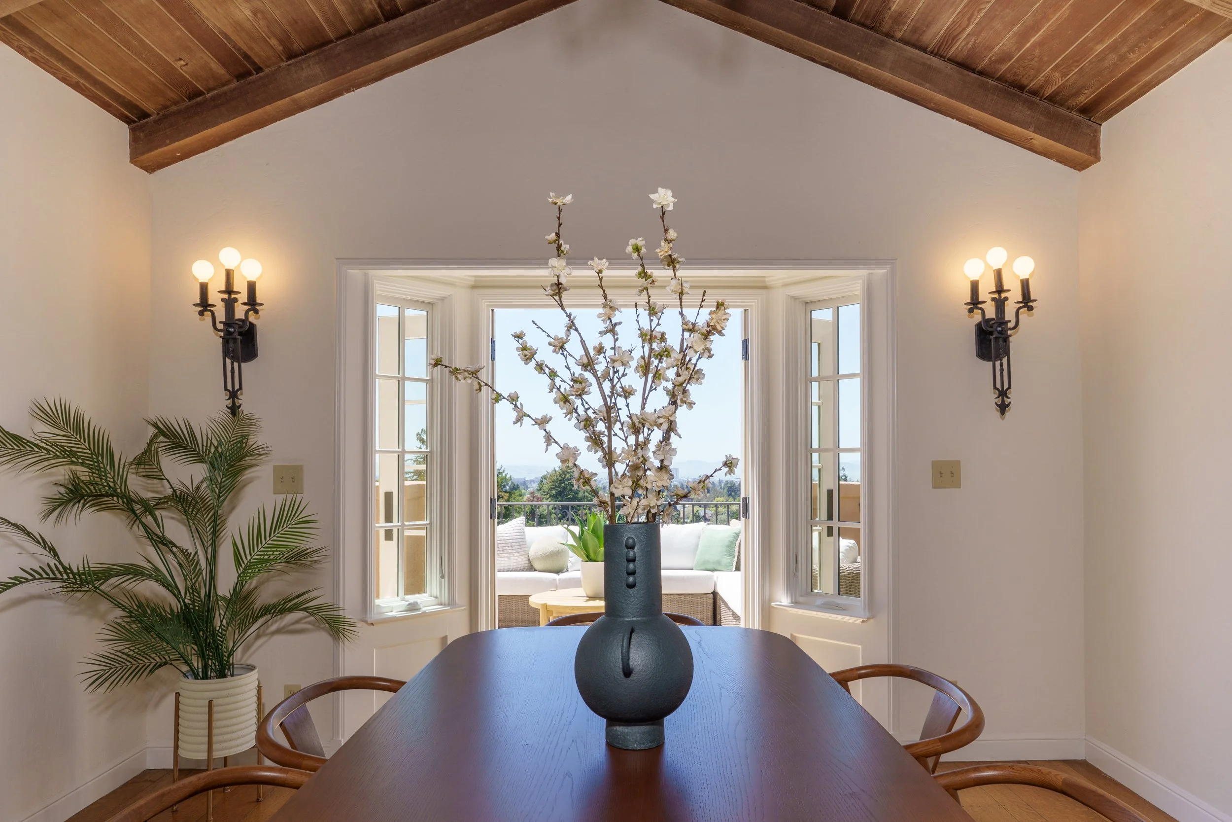 Interior of a dining room with a dark wood table, a large black vase with white flowering branches, and a window opening to a balcony with outdoor furniture and trees in the distance.