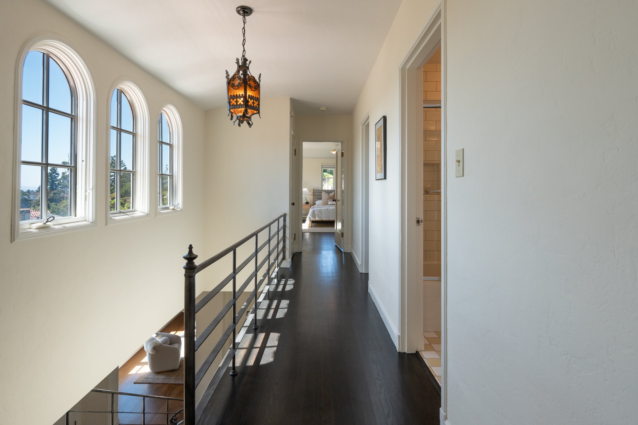 A hallway view inside a house with arched windows, dark hardwood flooring, white walls, a decorative hanging light fixture, and a bedroom visible at the end of the hall.