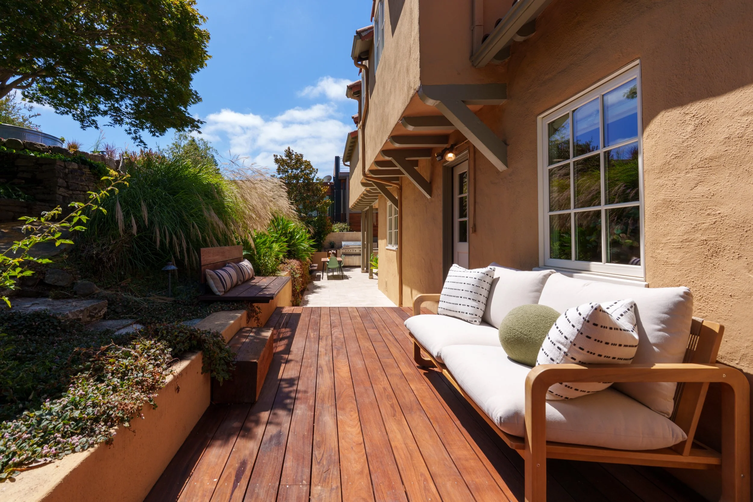 A backyard patio area with wooden decking, a white cushioned outdoor sofa with black and white pillows, a green round pillow, and lush green plants along a peach-colored wall of a house. There are large windows on the house, a small dining table with