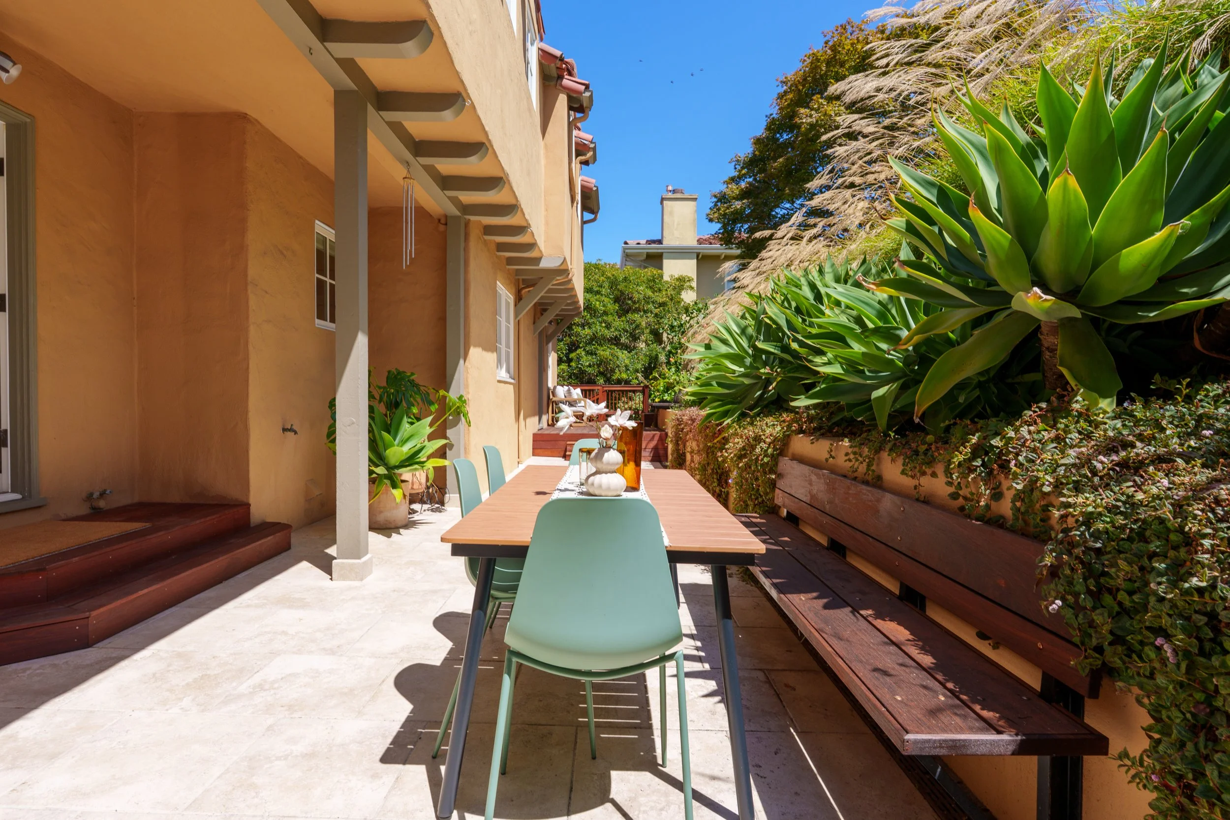 An outdoor patio area with a beige stucco house, a wooden dining table with mint green chairs, potted plants, and large green plants on the right side, under a pergola with wind chimes, with a view of neighboring houses and trees under a clear blue s