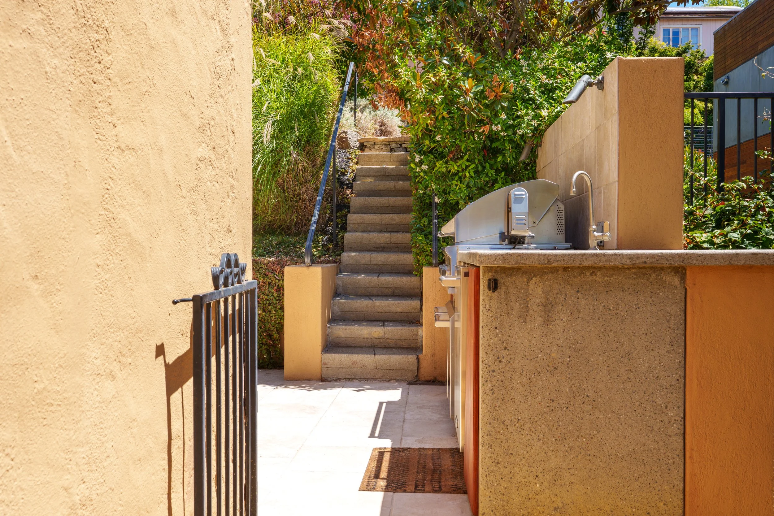 Outdoor scene of a staircase with concrete steps, greenery, and bushes leading upward, surrounded by beige walls and a yellow wall with a small outdoor sink, in bright sunlight.