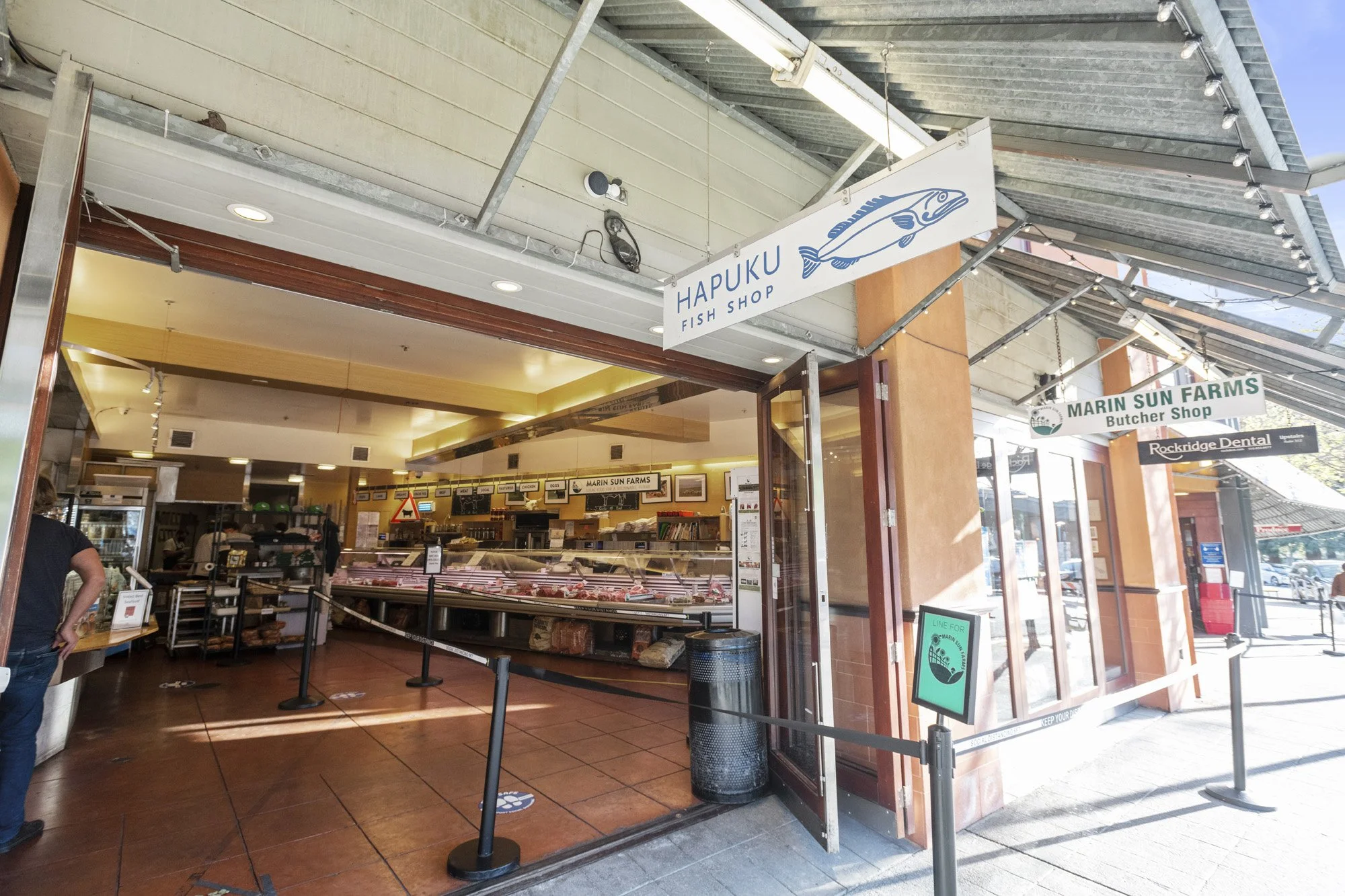 Fish shop with an open storefront, displaying fresh fish and seafood inside. Signs advertise 'Hapuku Fish Shop' and 'Marin Sun Farms Butcher Shop.' The shop has a modern design with indoor lighting and outdoor shading. There are social distancing mar