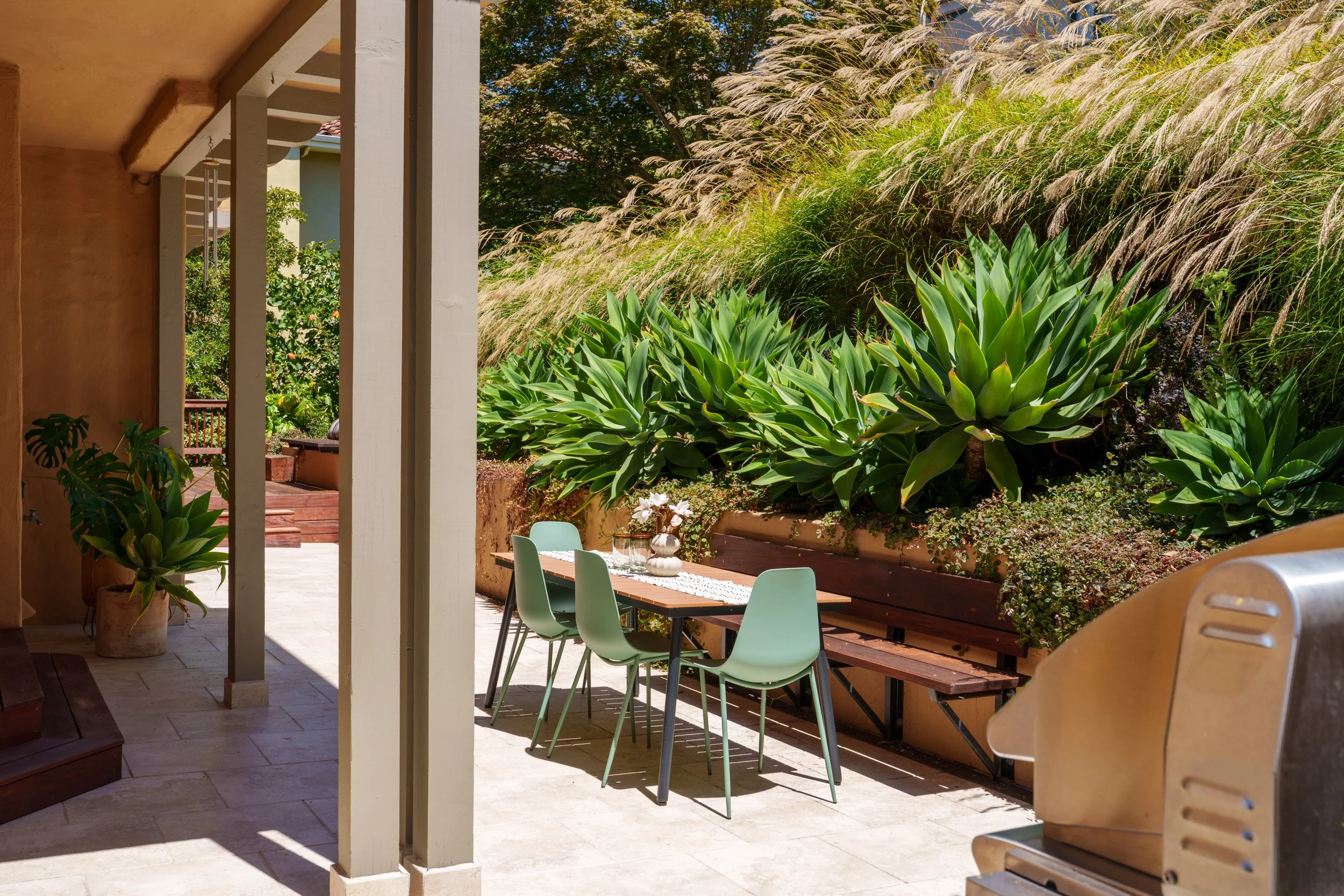 A backyard patio with a outdoor dining table and pastel green chairs, surrounded by lush green plants and tall ornamental grasses, with a tree in the background under a sunny sky.