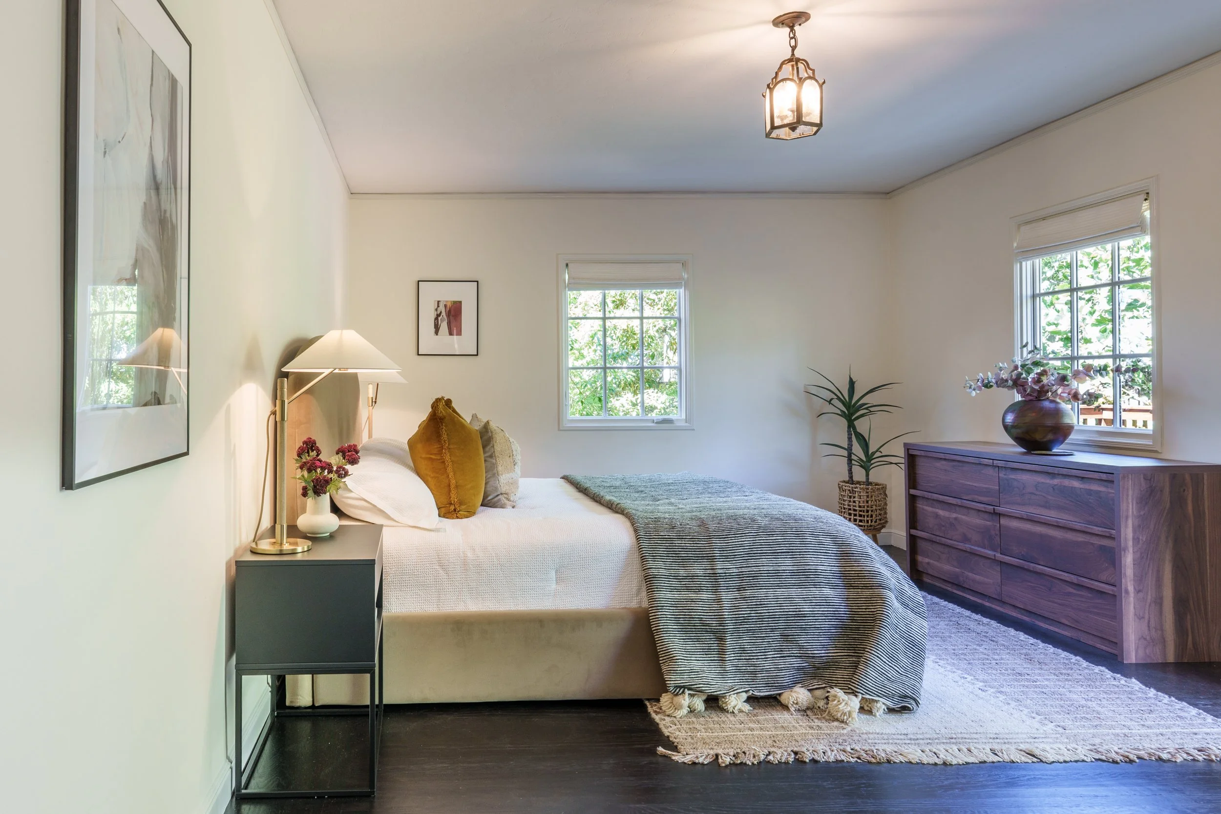 A bedroom with a bed, nightstand, and dresser, with large windows showing trees outside, and decorated with plants and artwork.