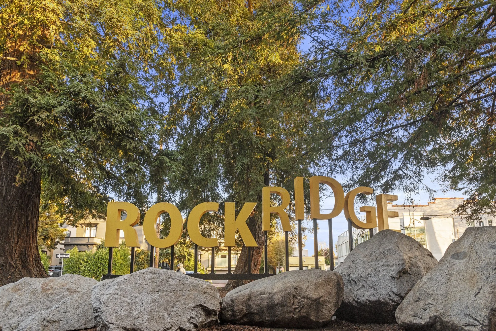 Rockridge sign in front of trees with yellow and green leaves, rocks in foreground, buildings and parked cars in background.