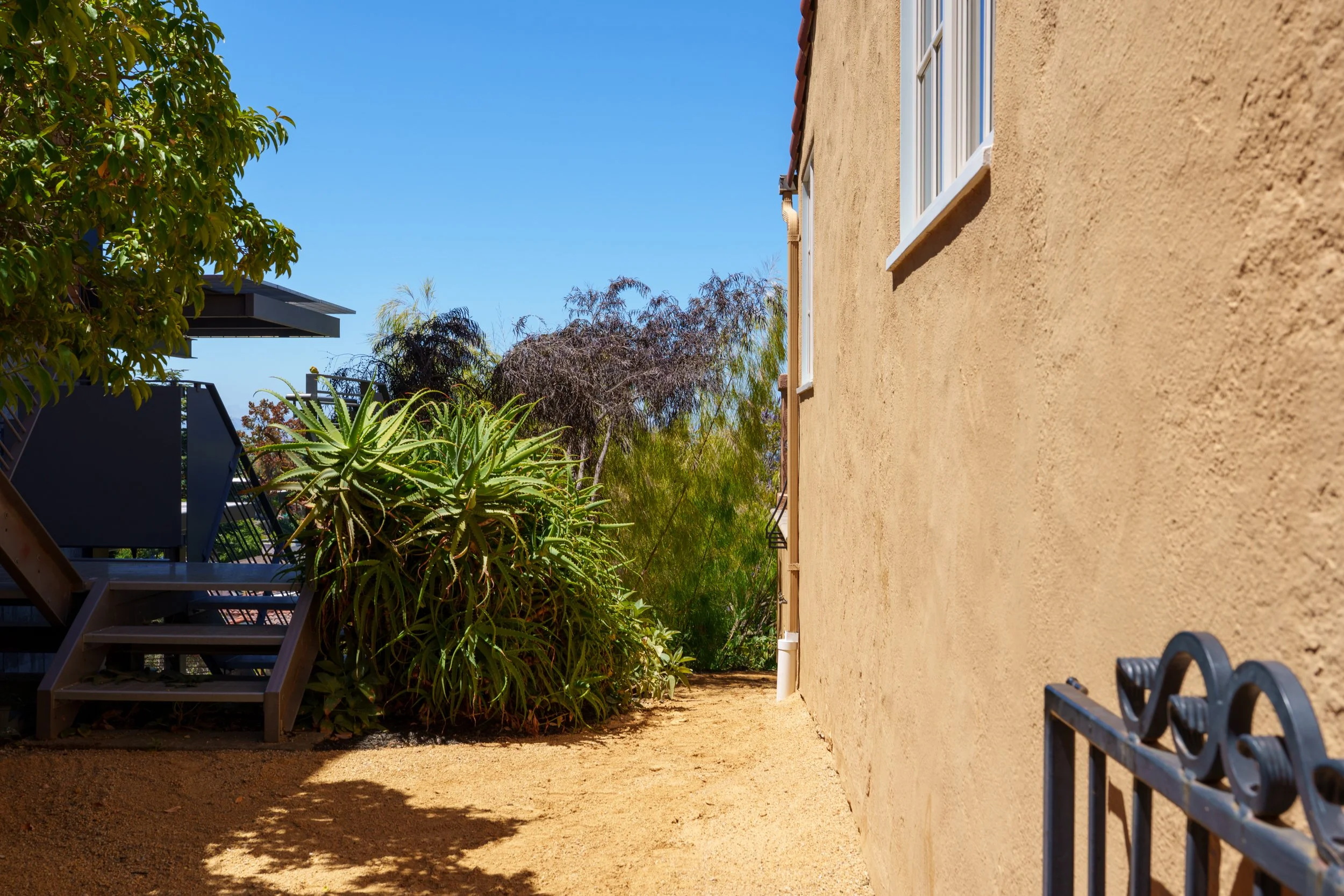 Side view of a beige stucco house with a window, a small deck with stairs, plants including a large aloe vera and trees, a clear blue sky, and a dirt yard.