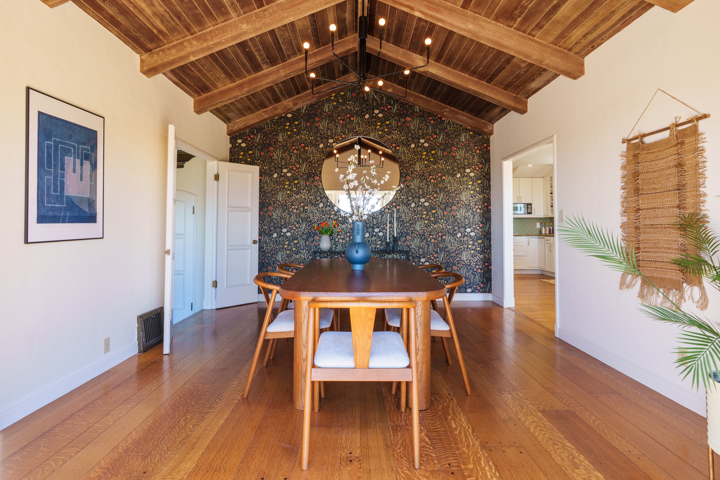 A dining room with a wooden table and chairs, a floral wallpaper wall with a round mirror, a black chandelier with exposed bulbs, and a decorative woven wall hanging. The room has a wooden ceiling and hardwood floors.