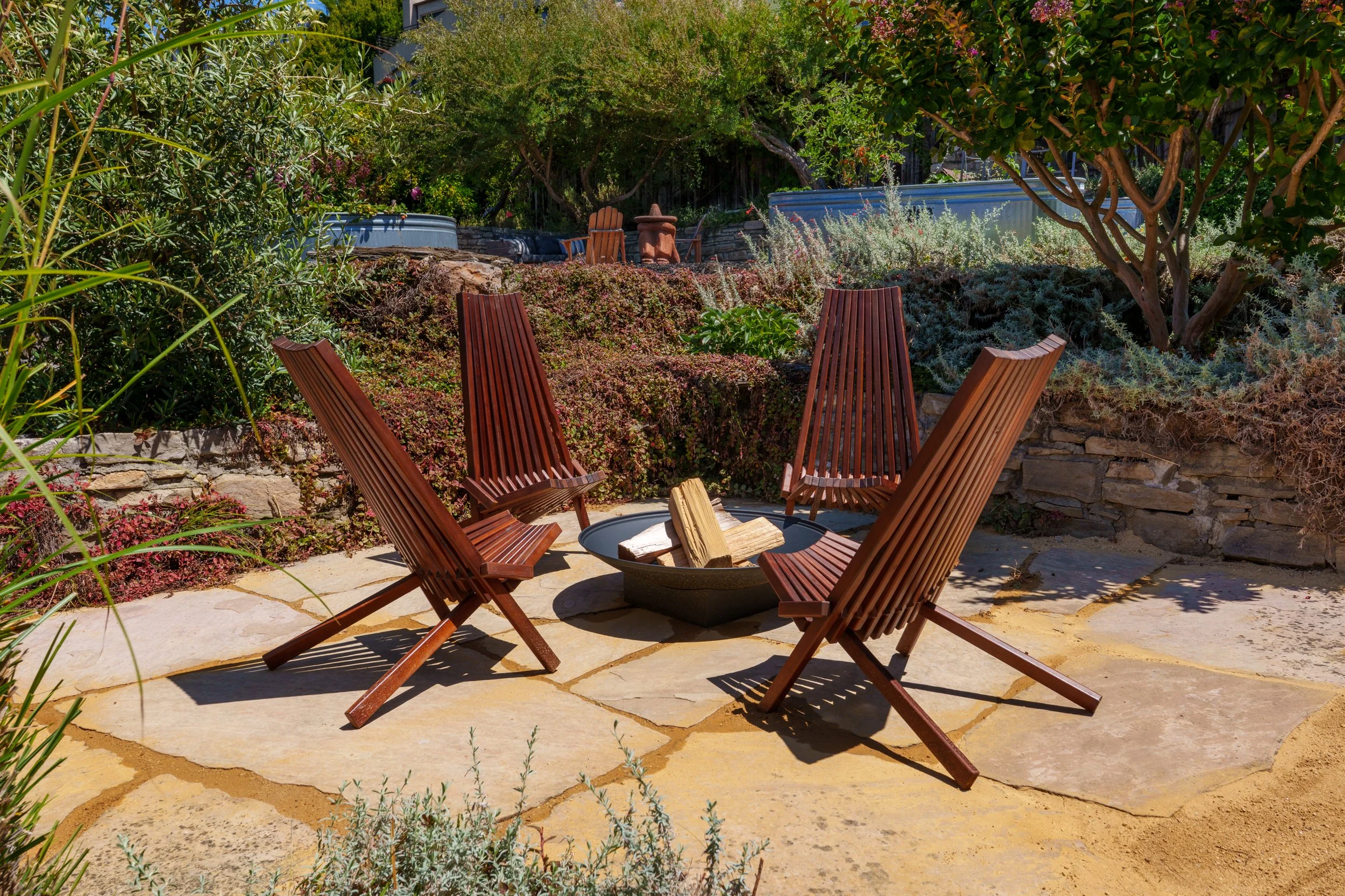 A sunny backyard garden with a fire pit surrounded by wooden chairs and lush greenery.