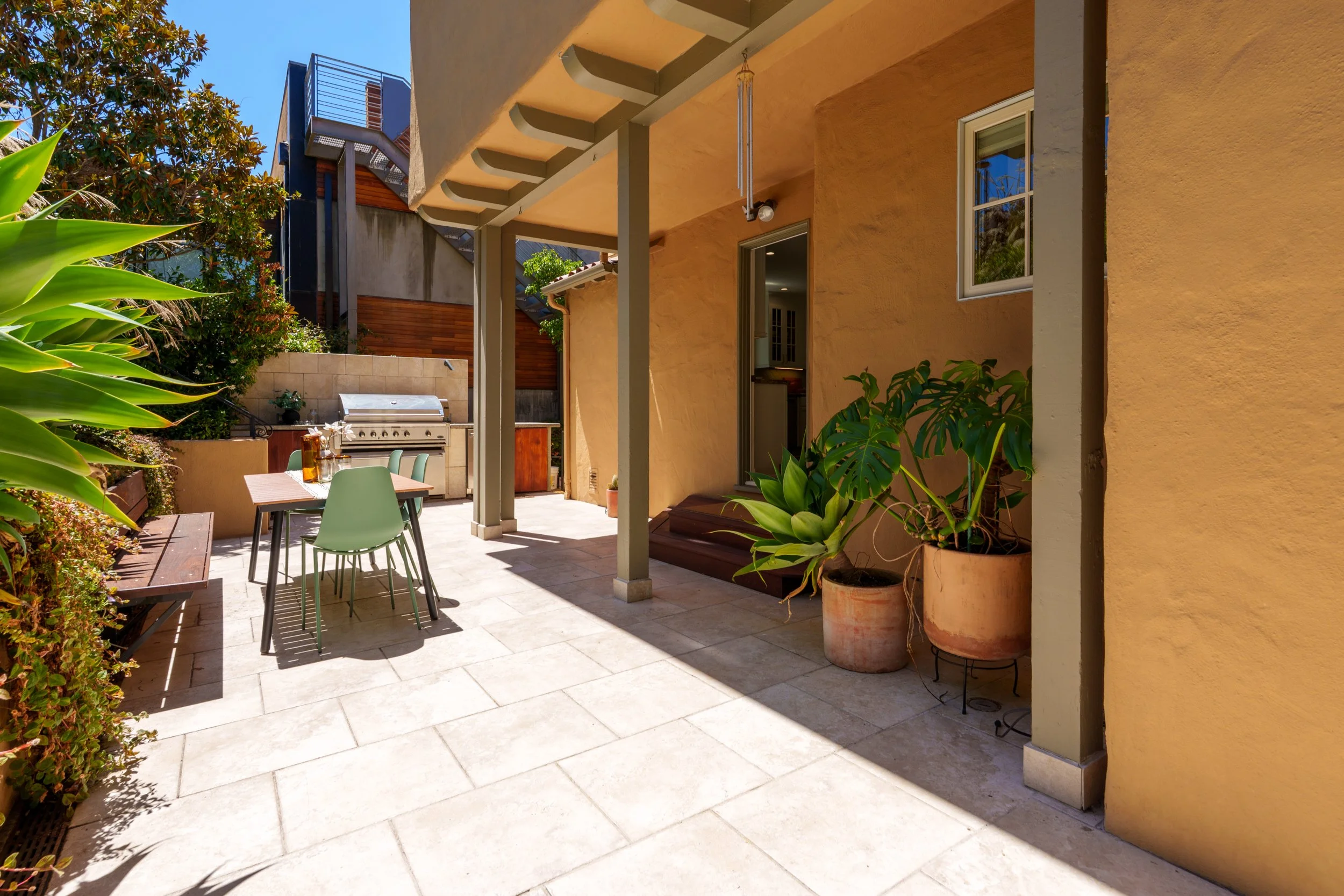 A backyard patio with outdoor furniture, a barbecue grill, lush plants, and a yellow stucco wall under a covered patio area.