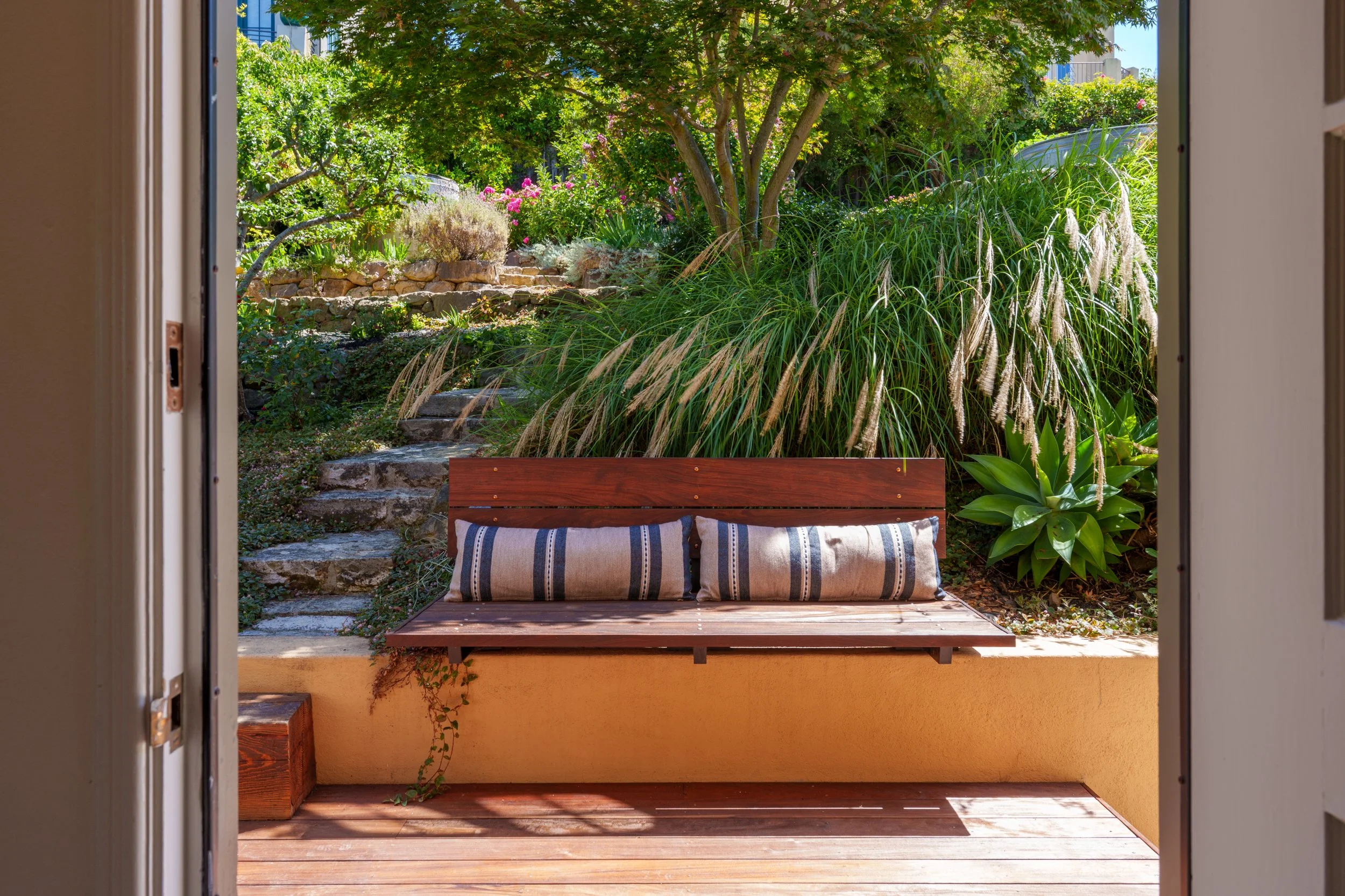 A view of a garden sitting area seen through an open door. The garden has a wooden bench with striped cushions, large leafy plants, and a variety of flowers and trees, with stairs leading up to greenery in the background.