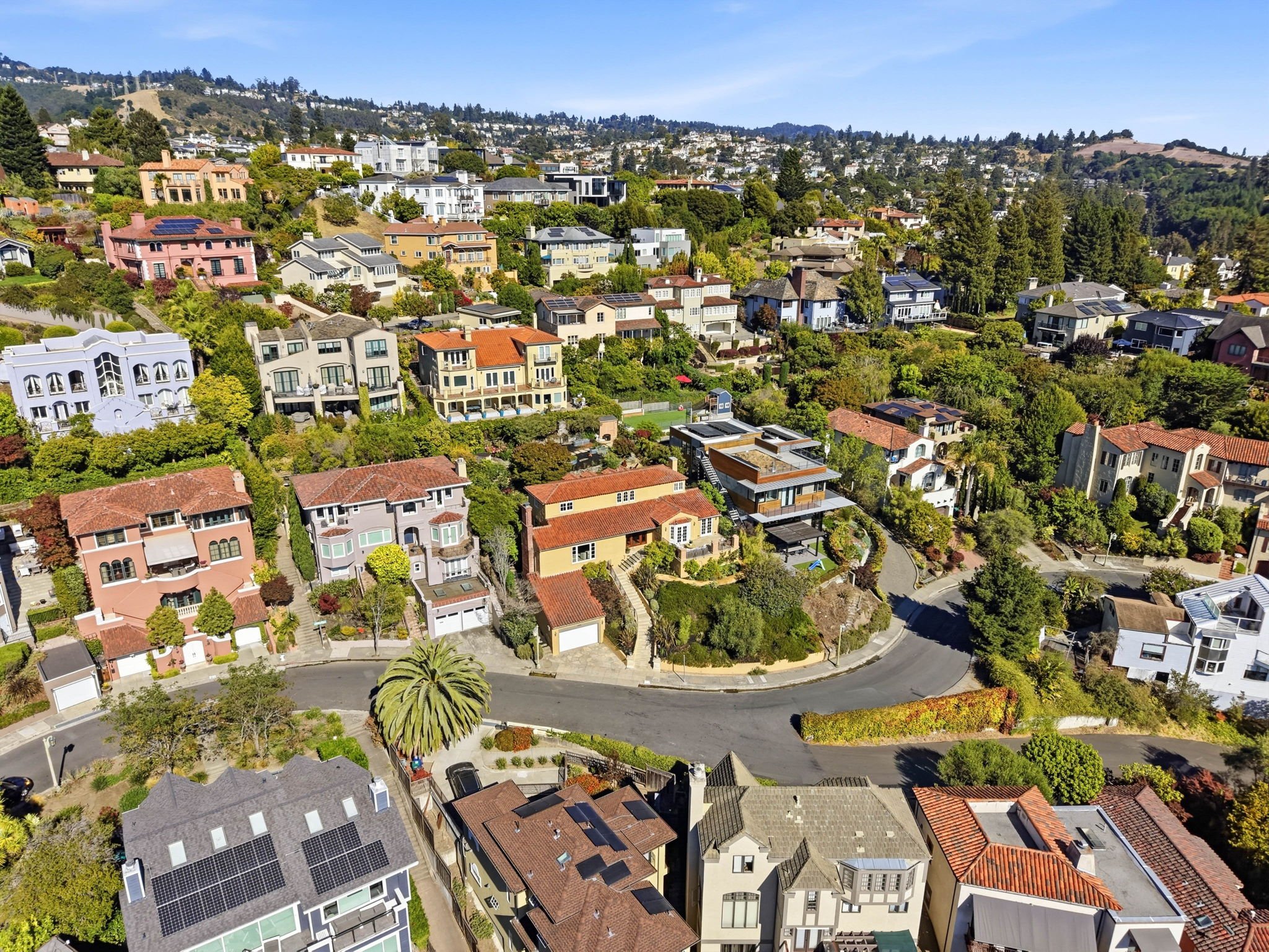 Aerial view of a hillside neighborhood with colorful houses and lush greenery with trees and bushes, under a clear blue sky.