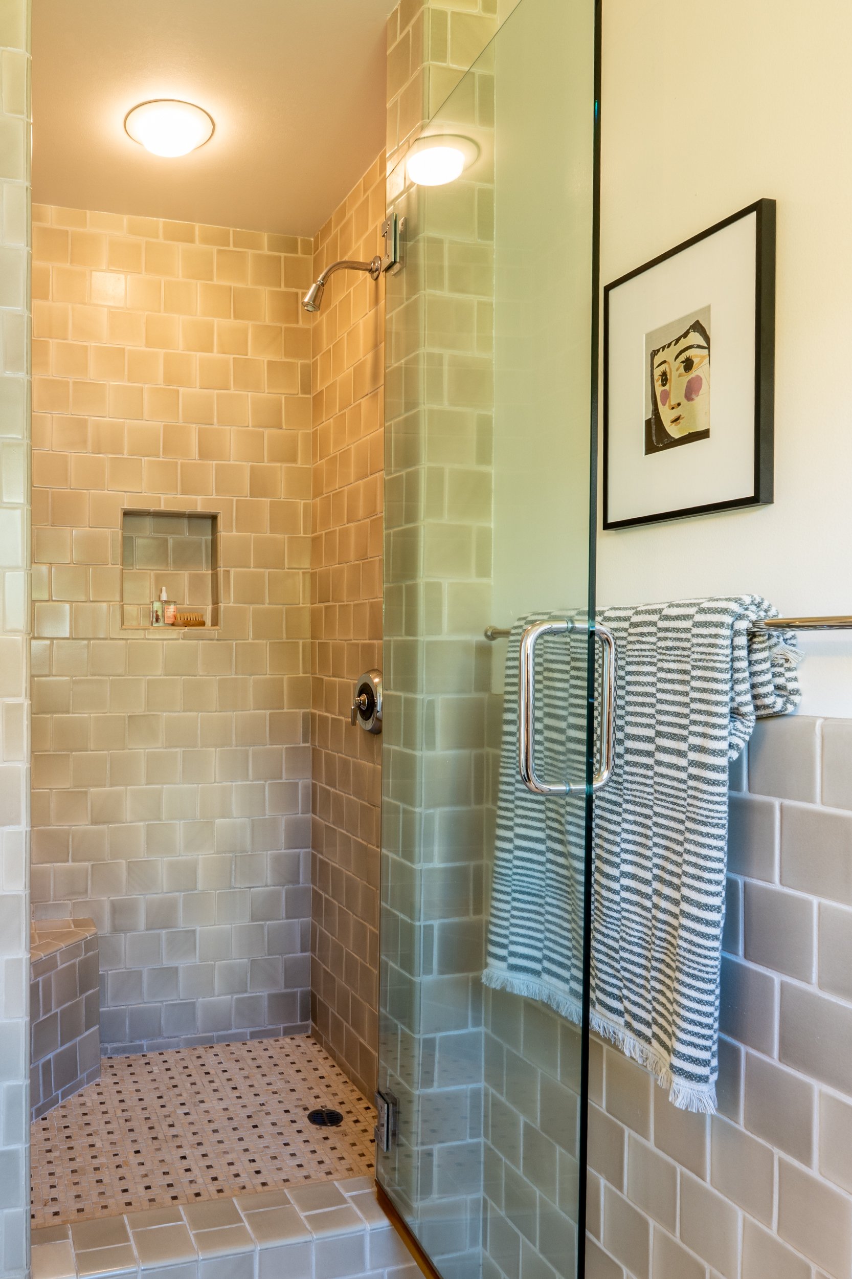 A modern walk-in shower with beige subway tiles, a built-in shelf, and a glass door. There is a black and white striped towel hanging on a towel bar and a framed artwork of a face on the wall outside the shower. The floor inside the shower has small 