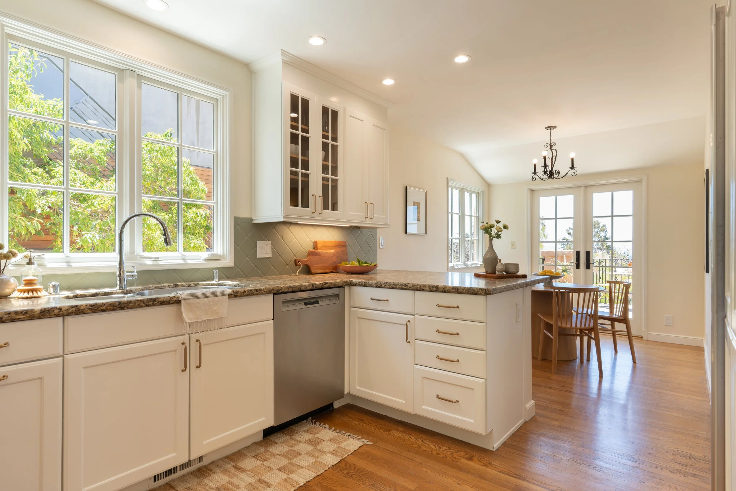Bright kitchen with white cabinets, granite countertops, large windows, and hardwood floors. There is a chandelier hanging above the dining area with a wooden table and chairs, and a vase with flowers on the table.