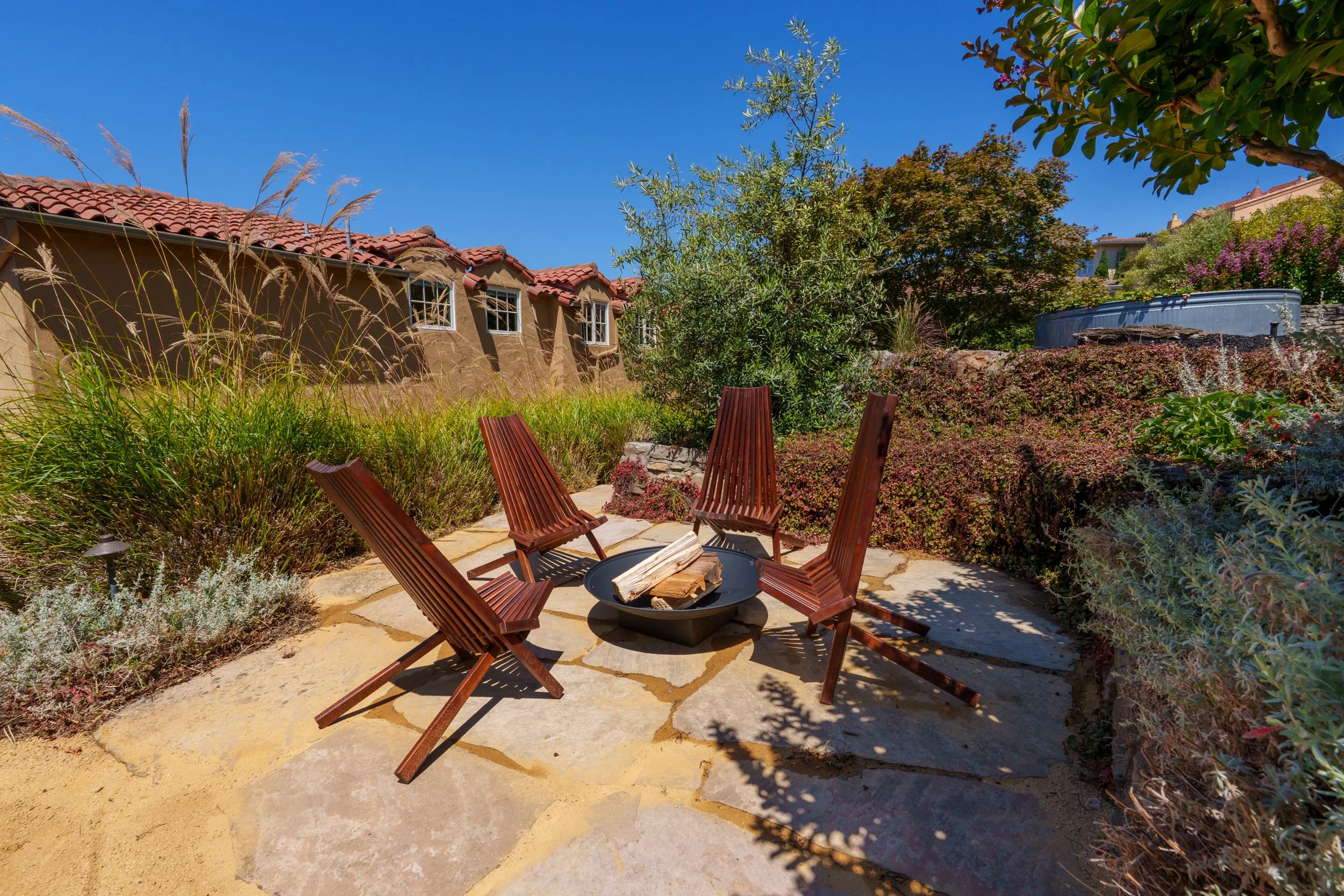 Outdoor patio with six wooden chairs arranged around a fire pit with logs, surrounded by lush green plants and trees, against the backdrop of a house with a red-tile roof and clear blue sky.
