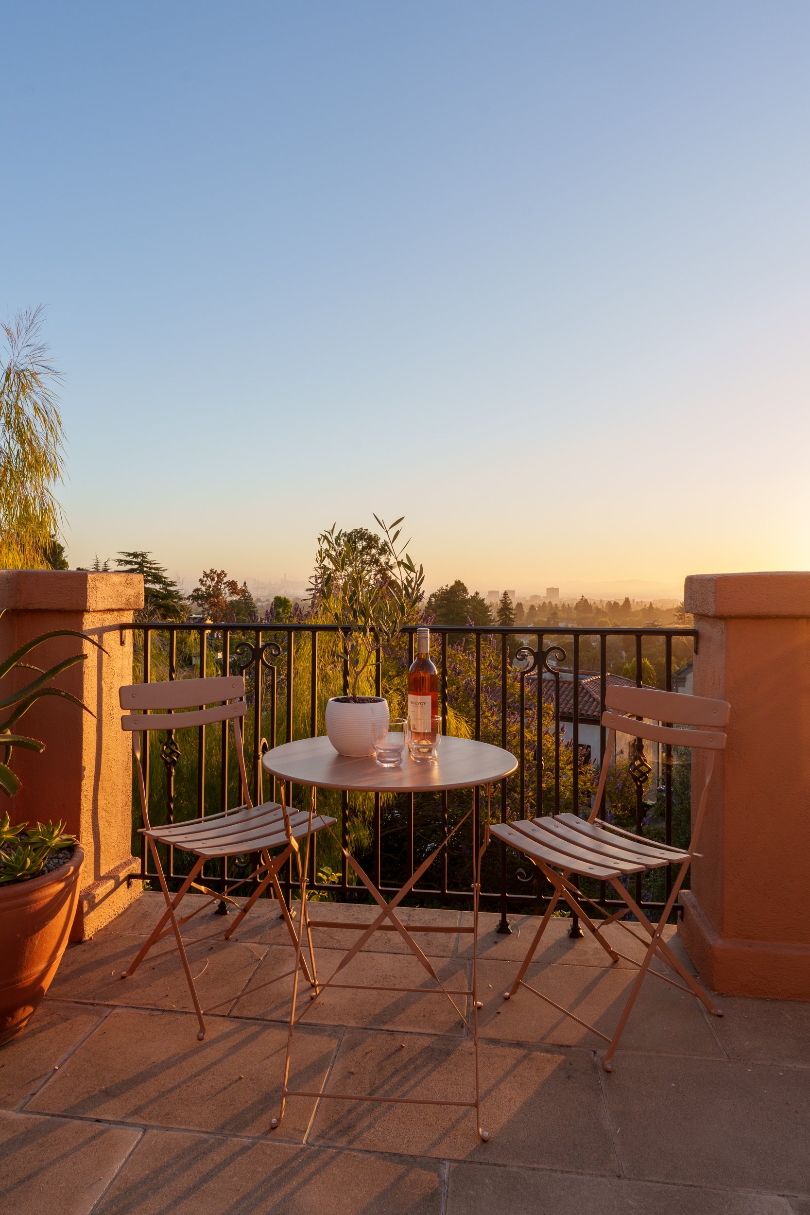 View from a balcony at sunset with a small round table, two chairs, a bottle of wine, a glass, and a plant, overlooking a landscape with trees and a hazy sky.