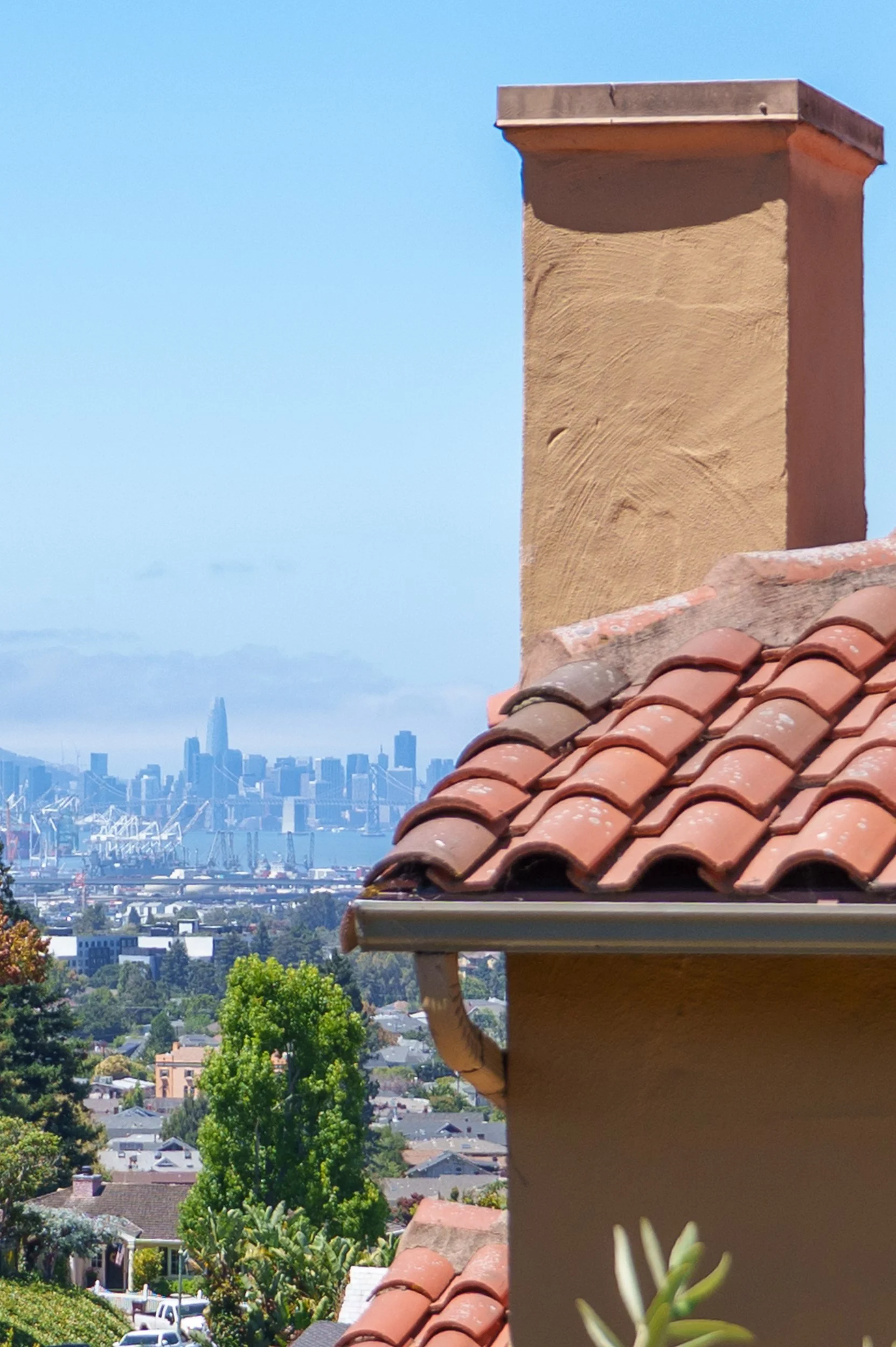 View of San Francisco skyline with tall buildings and the Bay Bridge, seen from a neighborhood with houses, trees, and a chimney in the foreground.