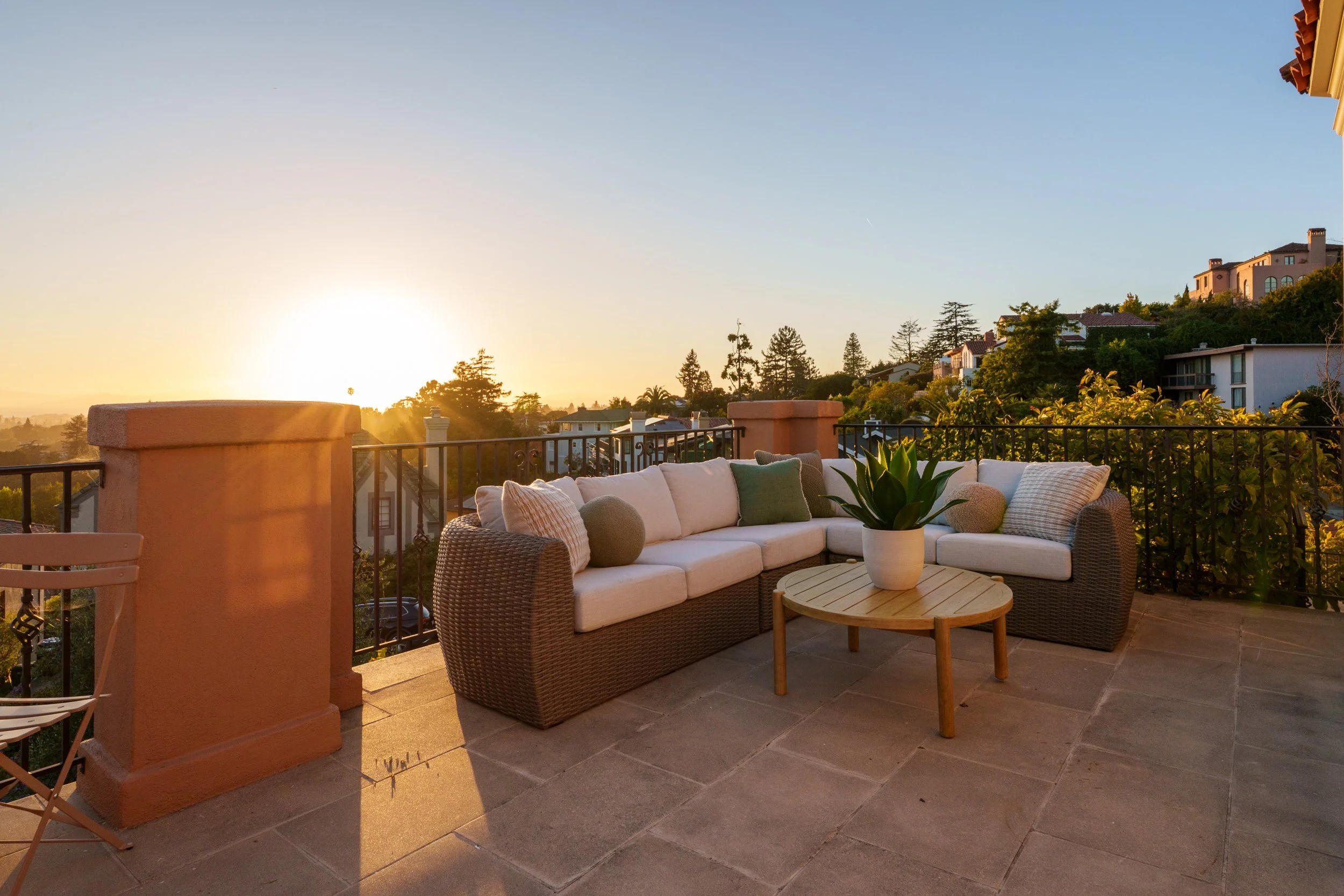 A rooftop balcony patio with outdoor furniture including a sectional sofa with cushions, a small round wooden coffee table, and a potted plant. The view features trees and houses on a hillside with the sun setting in the background.