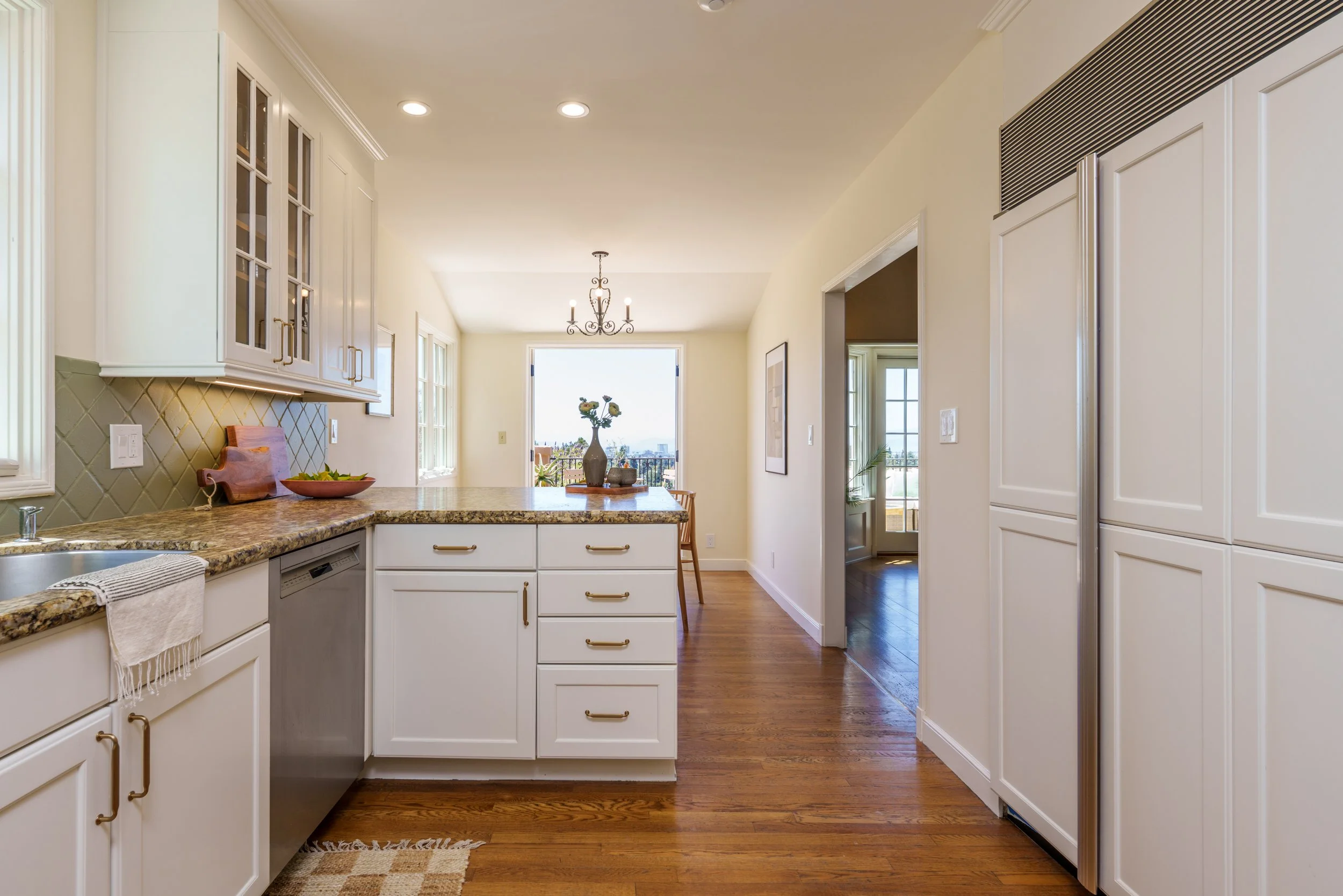 A bright kitchen with white cabinets, a kitchen island with a granite countertop, hardwood floors, a chandelier, and a view of the outdoors through a large window and a door.