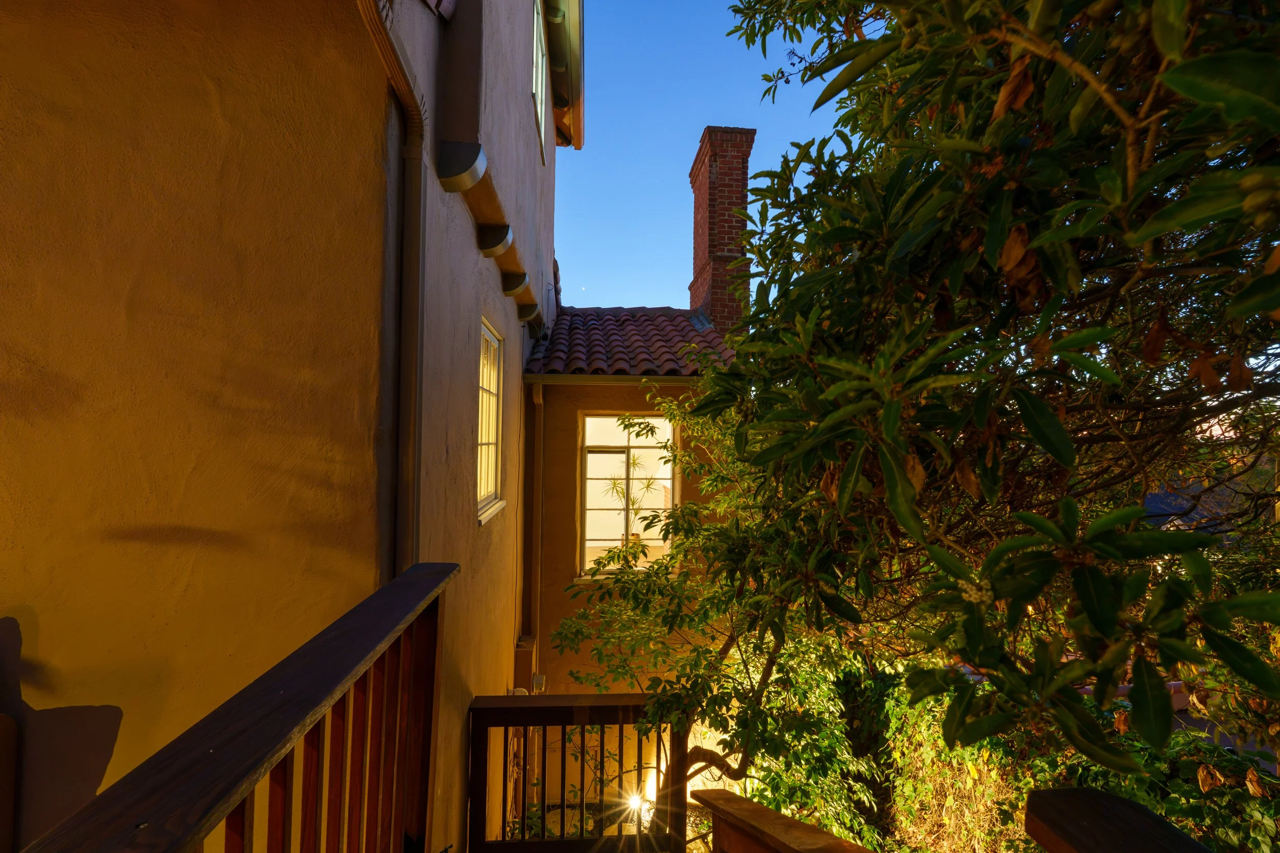 Outdoor porch area with yellow stucco wall, window, and wooden railing. A tall brick chimney extends above a roof with red tiles. Trees with green leaves partially obscure the view, and the sky is clear blue with a visible moon or planet. Warm lighti