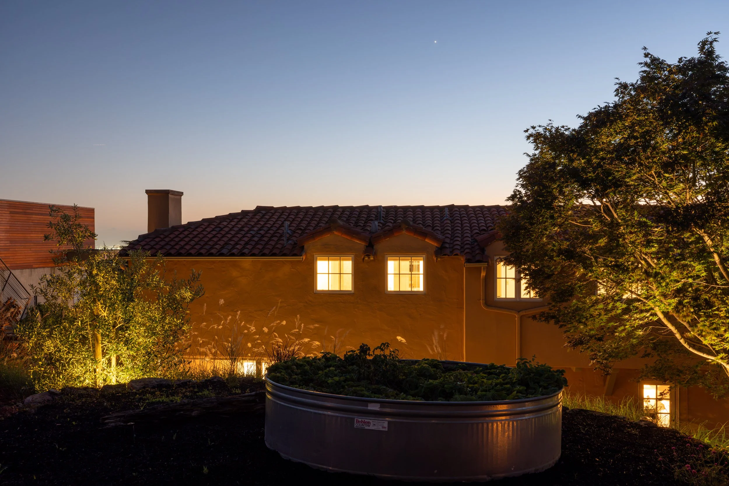 Residential house with illuminated windows under a clear evening sky, trees in the foreground, and a large planter with plants.