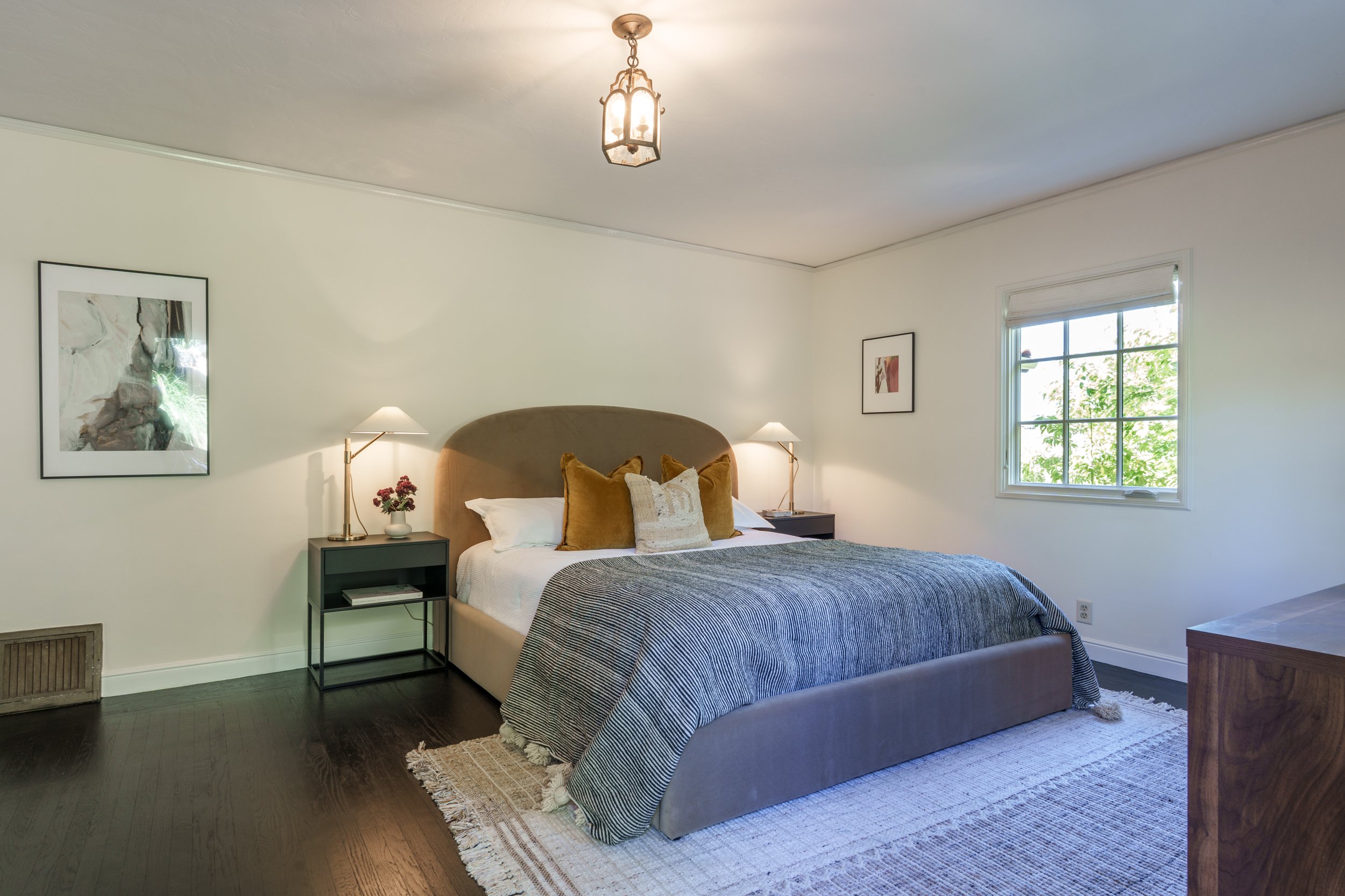 A bedroom with a bed, two side tables with lamps, and artwork on the white walls. There is a window with a view of greenery outside.